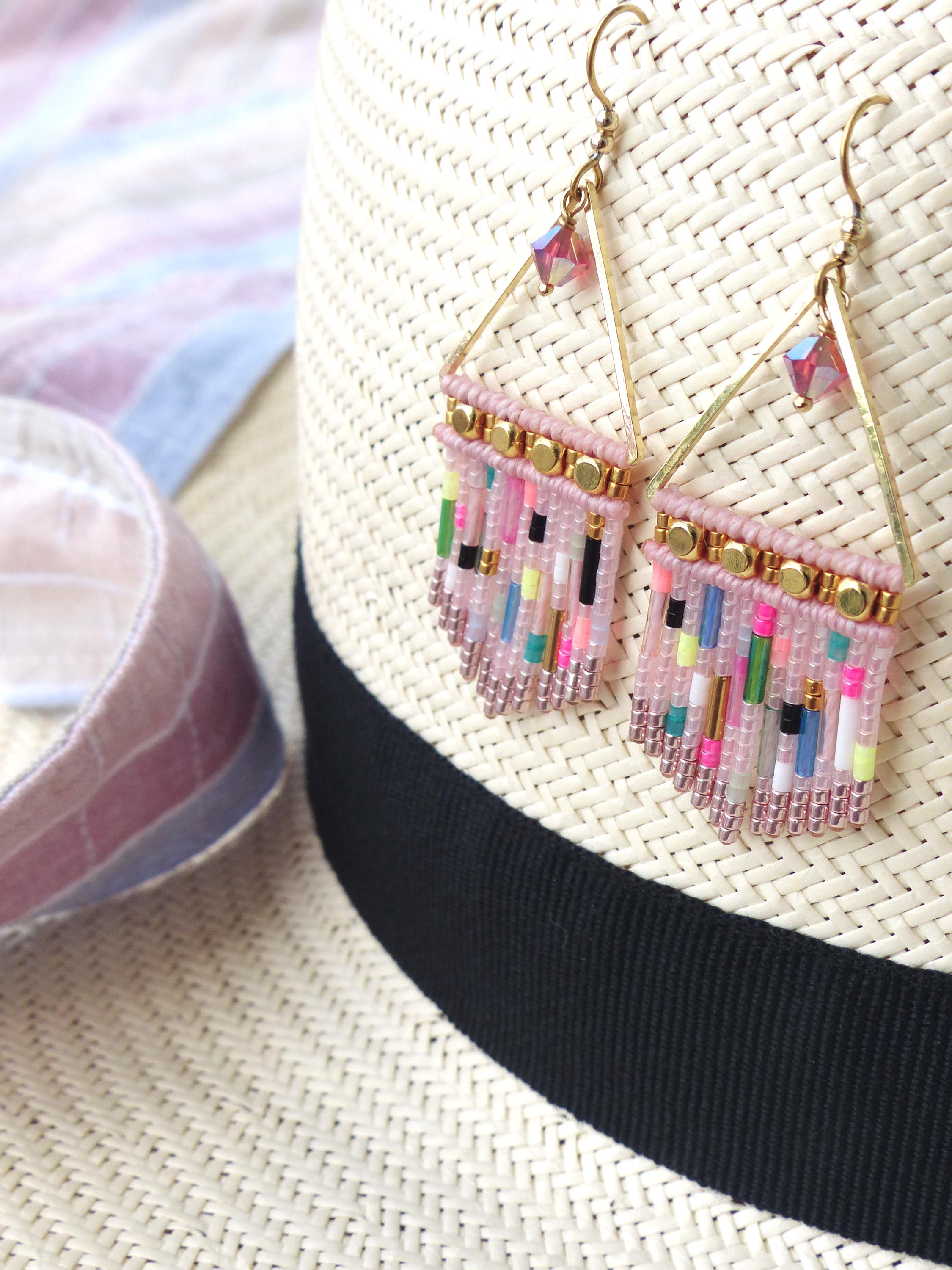 Peut inclure: Boucles d'oreilles faites à la main avec un design géométrique. Les boucles d'oreilles présentent une base rose avec des perles colorées dans les tons de rose, vert, bleu et jaune. Elles sont suspendues à des cadres triangulaires dorés et sont présentées sur un chapeau de paille.