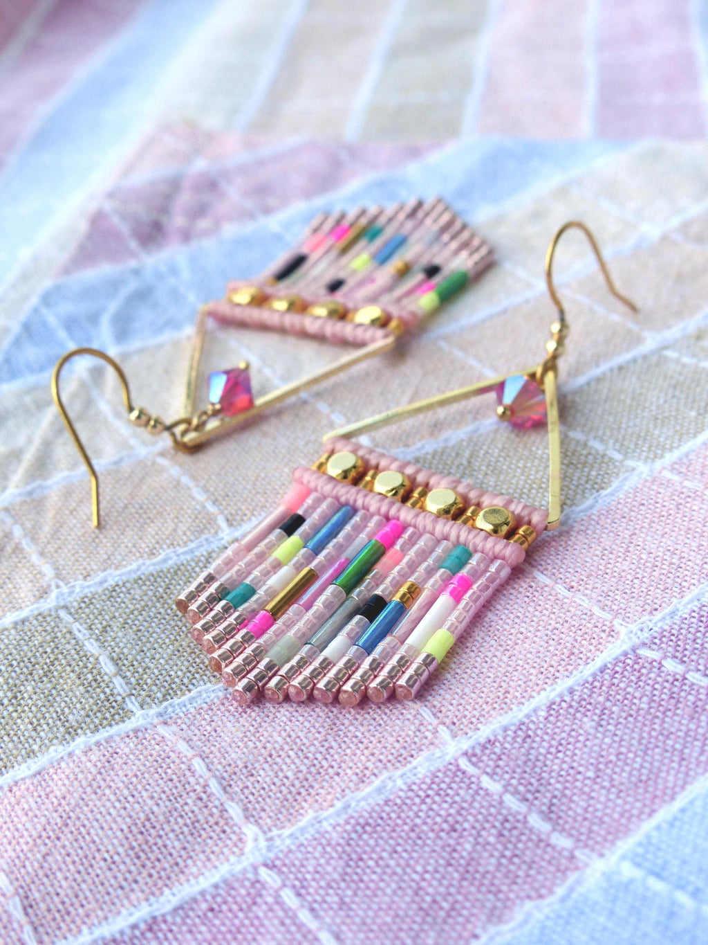 Peut inclure: Une paire de boucles d'oreilles faites à la main avec un design de franges perlées roses. Les boucles d'oreilles ont des crochets d'oreilles dorés et un cadre géométrique. La frange est ornée de perles colorées dans les tons de rose, vert, jaune et bleu. Les boucles d'oreilles sont présentées sur un tissu à motifs roses et blancs.