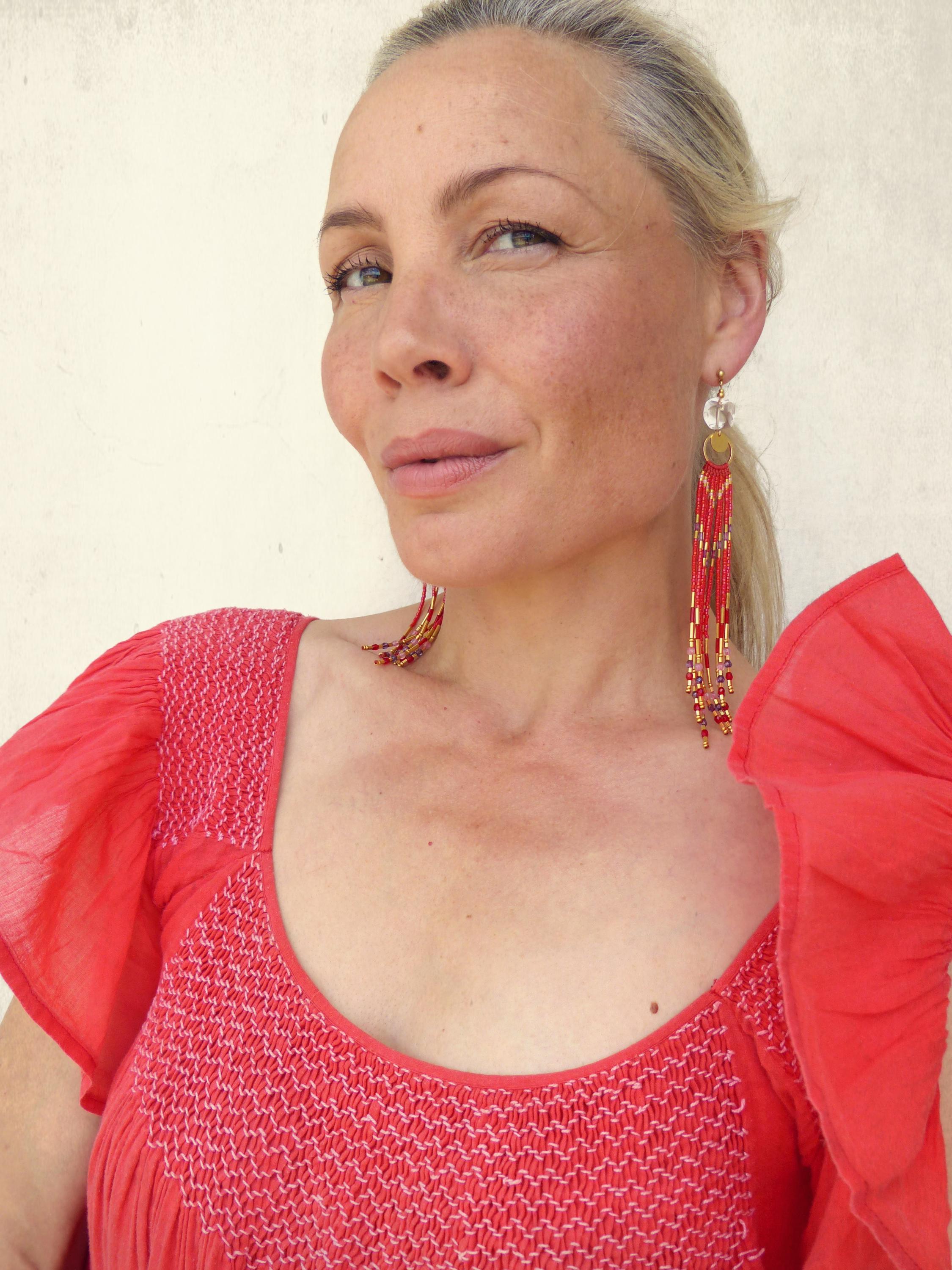 Peut inclure: Boucles d'oreilles pendantes rouges à perles avec des détails dorés. La femme porte un haut rouge à col rond et à manches à volants. Le haut présente un motif texturé sur le corsage.