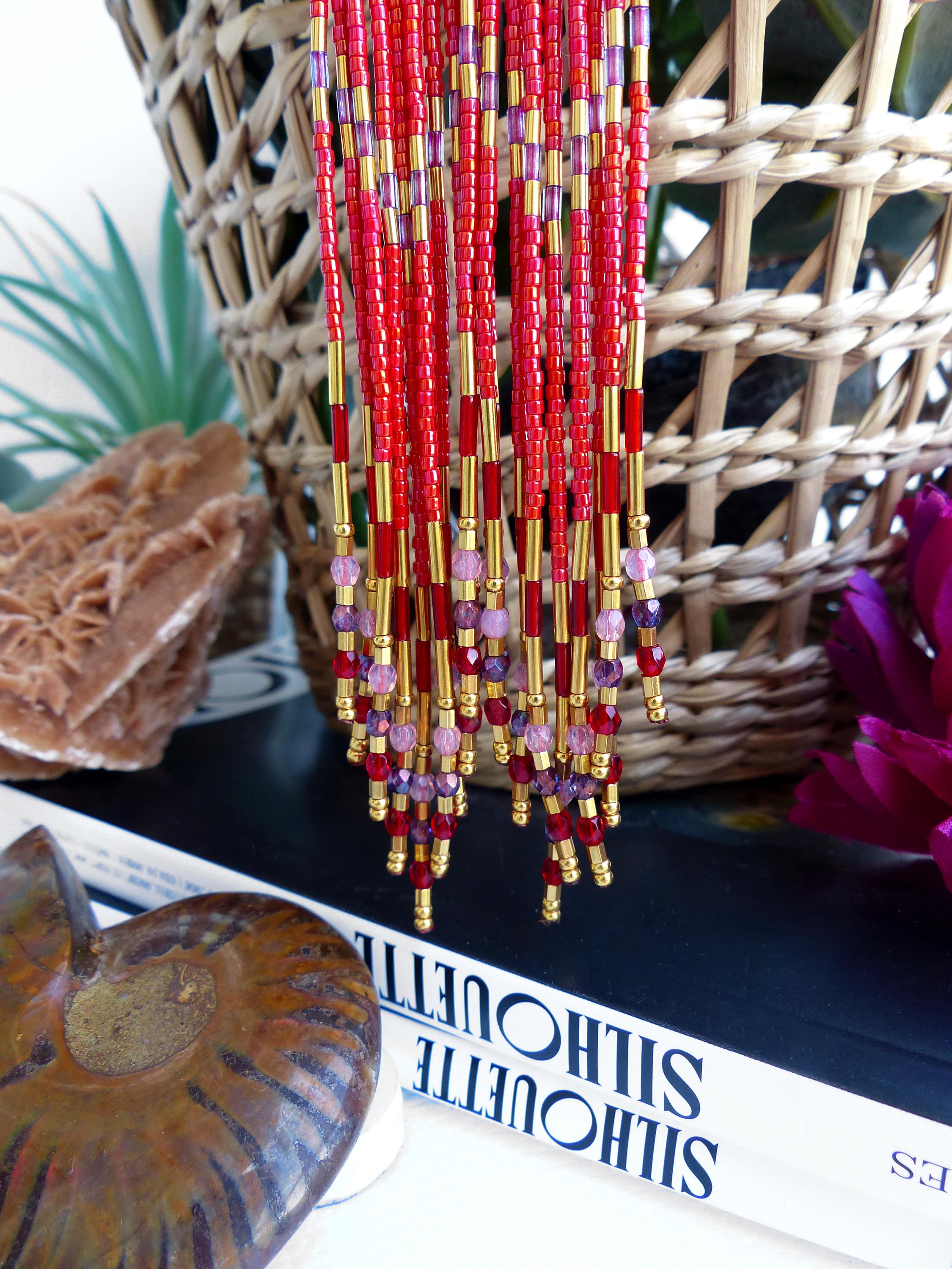 Peut inclure: Boucles d'oreilles en perles rouges avec des accents dorés et des perles violettes. Les boucles d'oreilles sont suspendues devant un panier en osier et un livre intitulé "Silhouette".