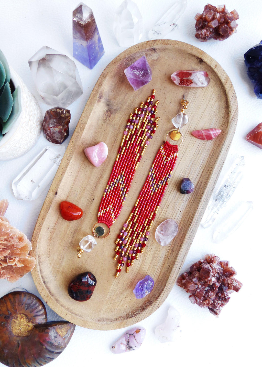 Peut inclure: Boucles d'oreilles rouges en perles avec des détails dorés et des gouttes de cristal de roche, présentées sur un plateau en bois avec des cristaux et des pierres précieuses. Les boucles d'oreilles ont un motif géométrique et une frange.