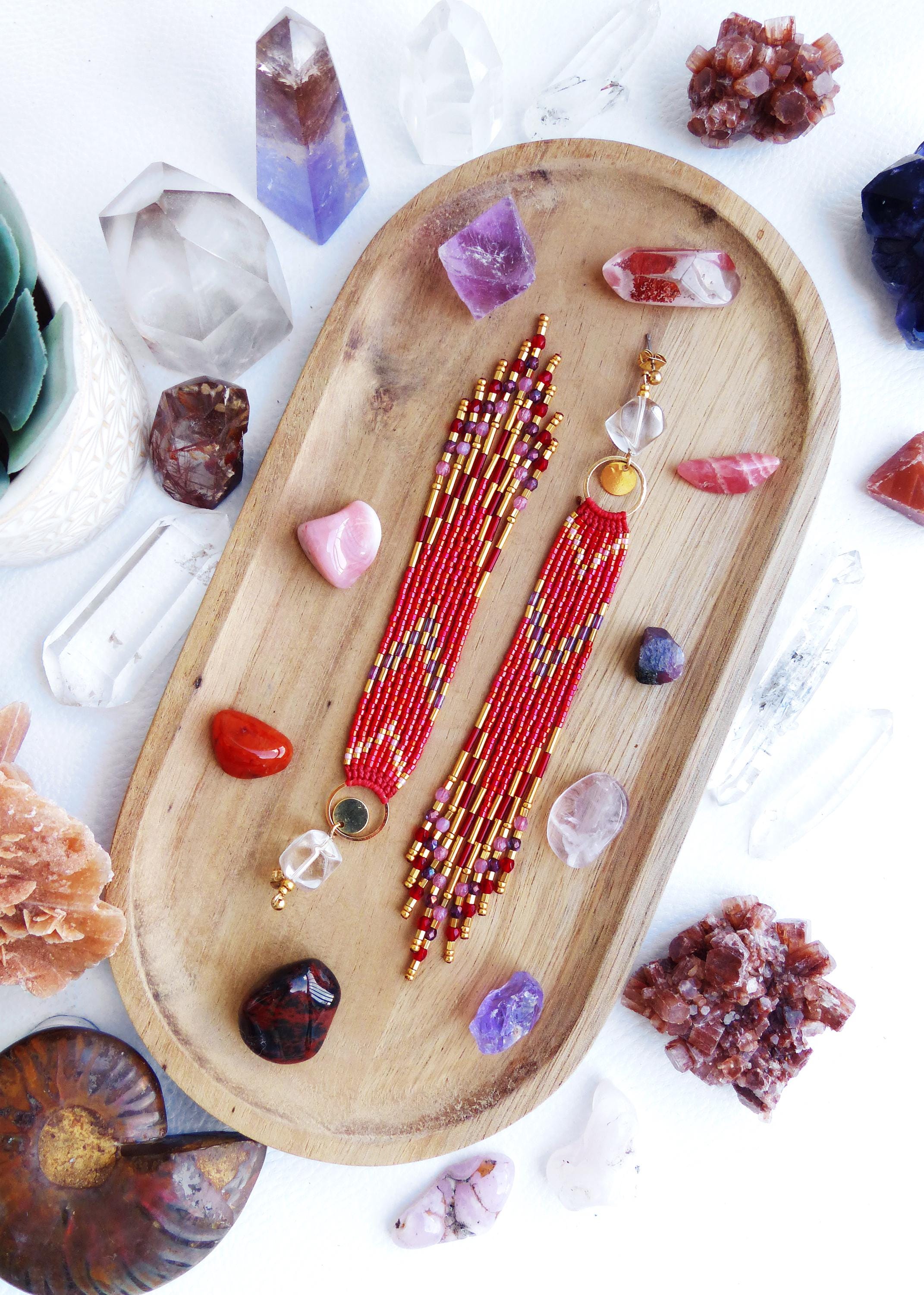 Peut inclure: Boucles d'oreilles rouges en perles avec des détails dorés et des gouttes de cristal de roche, présentées sur un plateau en bois avec des cristaux et des pierres précieuses. Les boucles d'oreilles ont un motif géométrique et une frange.