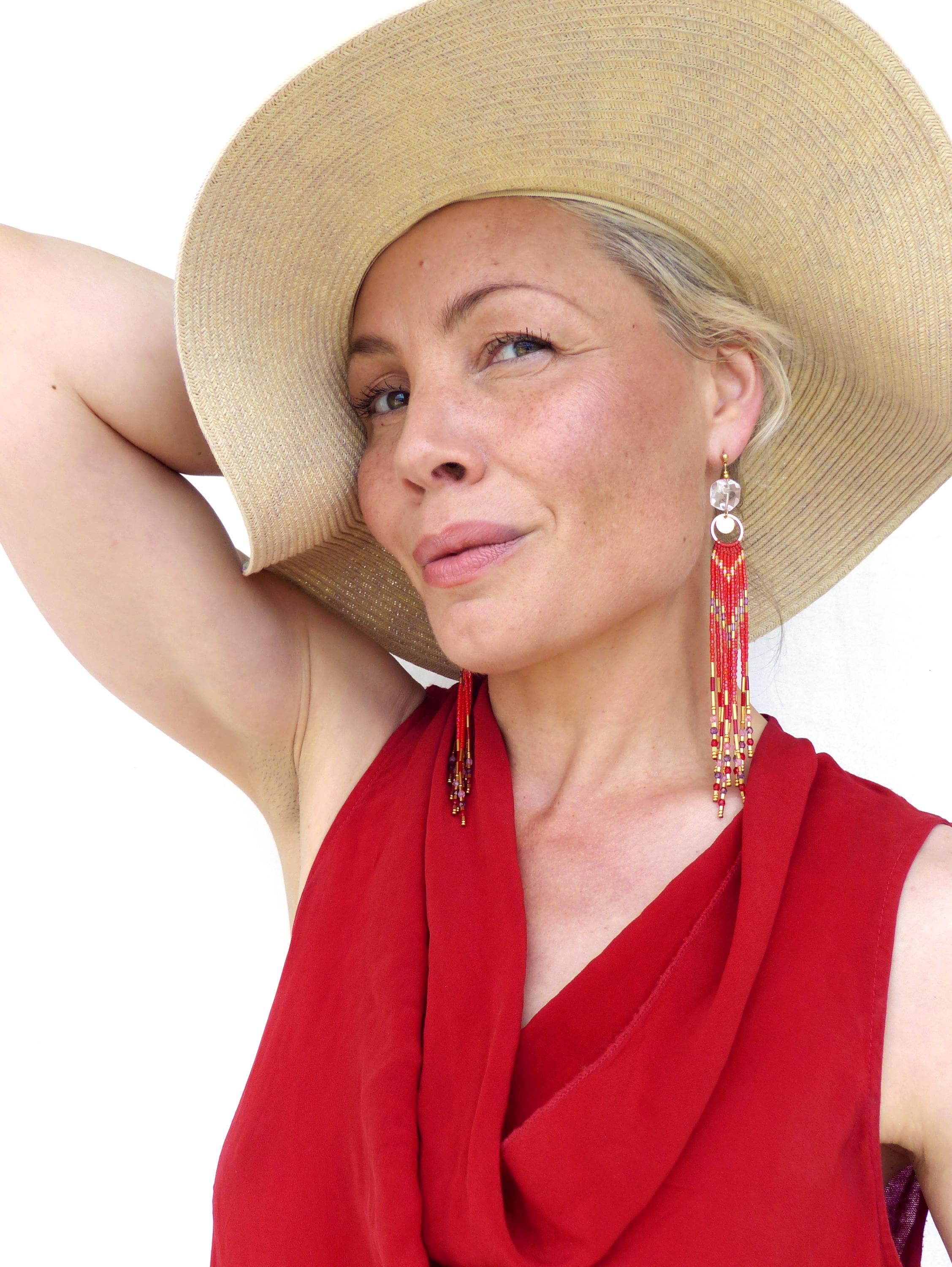 Peut inclure: Une femme porte un chapeau de paille à larges bords et une robe rouge. De longues boucles d'oreilles pendantes avec des perles rouges et dorées sont visibles. Les boucles d'oreilles ont une pièce supérieure transparente et rectangulaire. La femme regarde l'appareil photo.