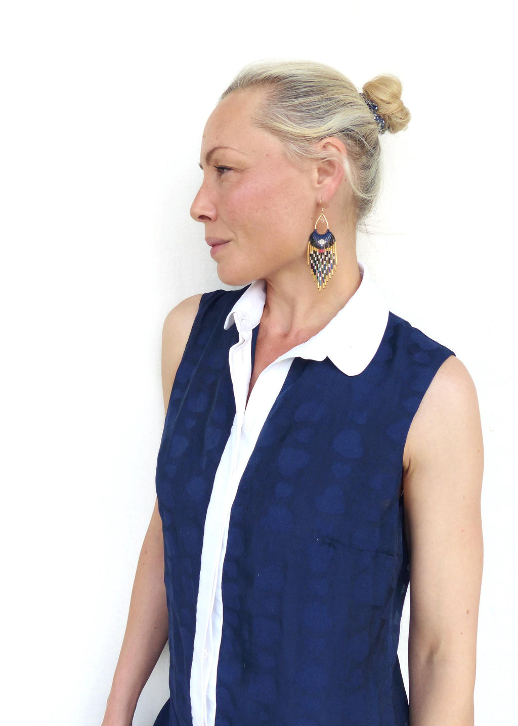 Peut inclure: Boucles d'oreilles géométriques en or, bleu marine et blanc. Les boucles d'oreilles ont une forme de goutte d'eau. Le modèle porte un haut sans manches bleu marine avec un col blanc. Bijoux.