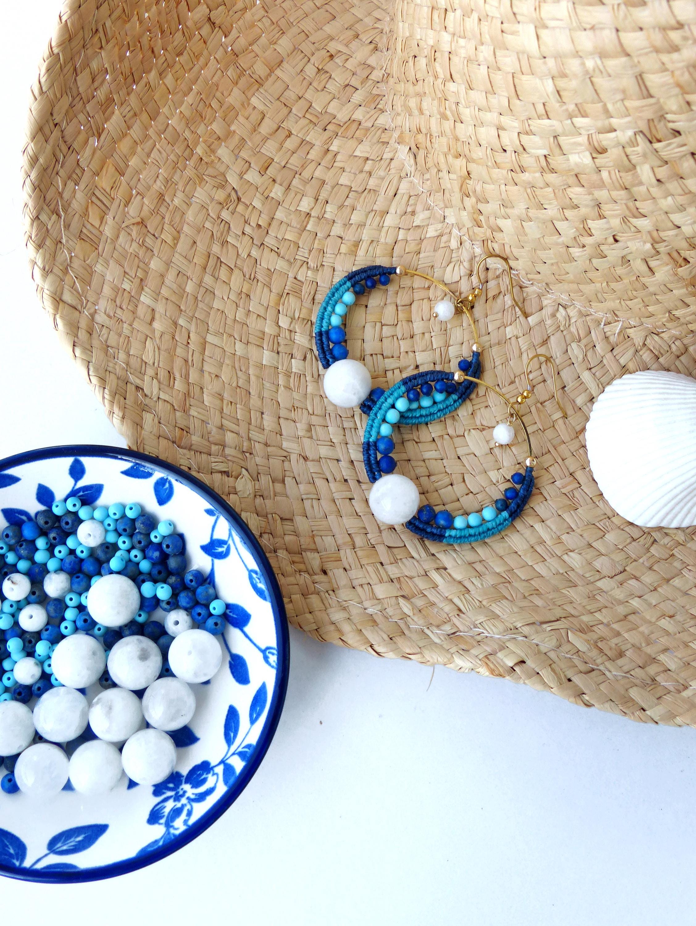 Peut inclure: Boucles d'oreilles créoles en macramé faites à la main, dans des tons de bleu et turquoise, ornées de perles blanches et d'une grosse sphère blanche. Elles reposent sur une surface en paille tressée, avec un petit plat de perles bleu et blanc.