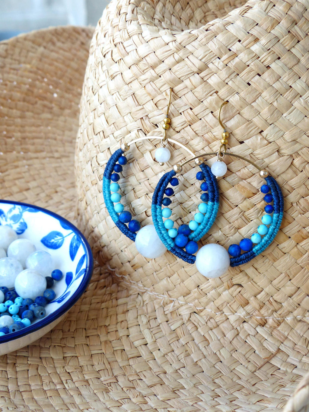 Peut inclure: Boucles d'oreilles créoles faites à la main sur le thème nautique. Les boucles d'oreilles sont composées d'anneaux en métal doré, enveloppés de fil bleu et turquoise, ornés de perles bleues et d'accents en pierre blanche. Présentées sur un chapeau de paille.