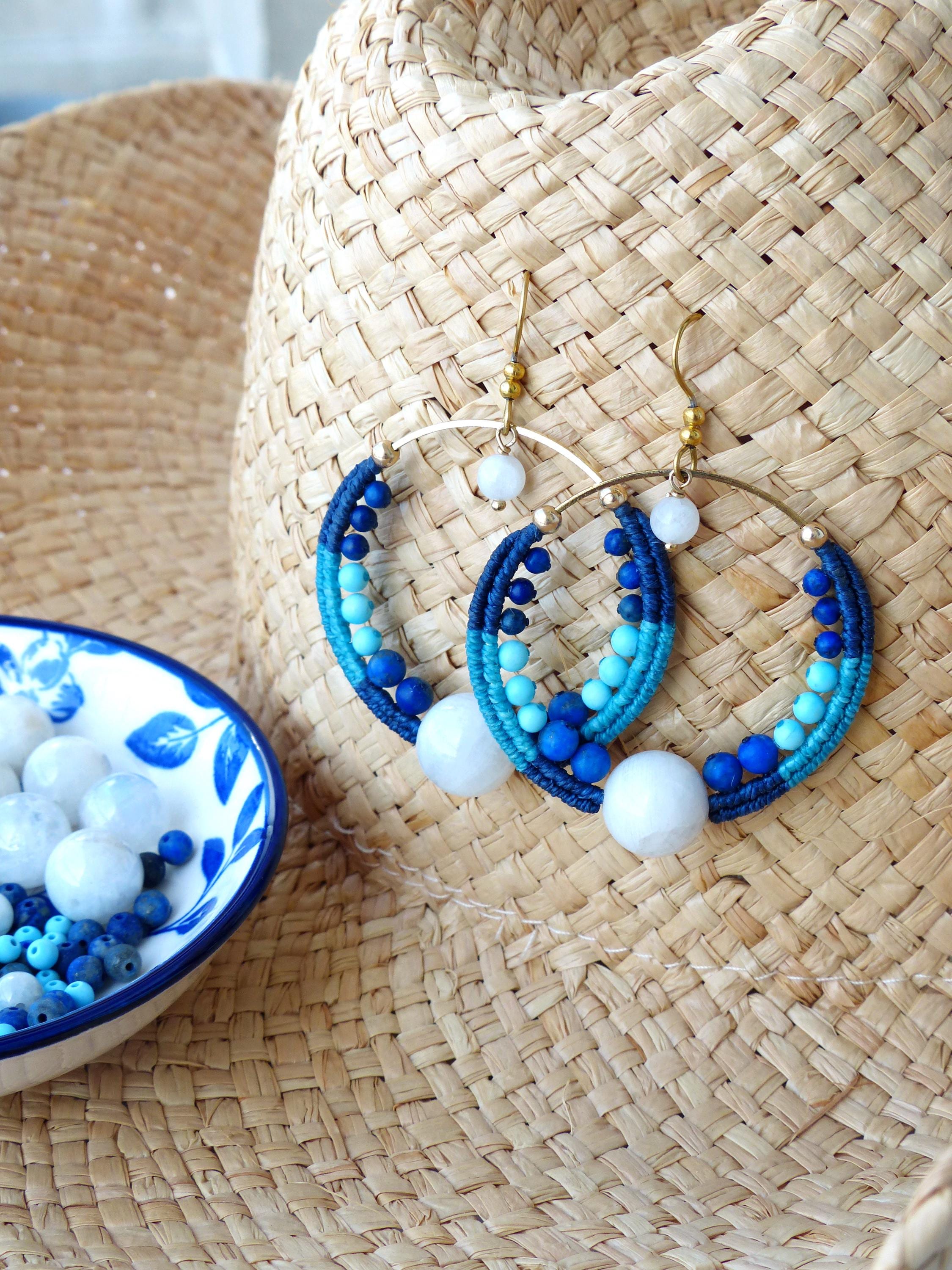 Peut inclure: Boucles d'oreilles créoles faites à la main sur le thème nautique. Les boucles d'oreilles sont composées d'anneaux en métal doré, enveloppés de fil bleu et turquoise, ornés de perles bleues et d'accents en pierre blanche. Présentées sur un chapeau de paille.