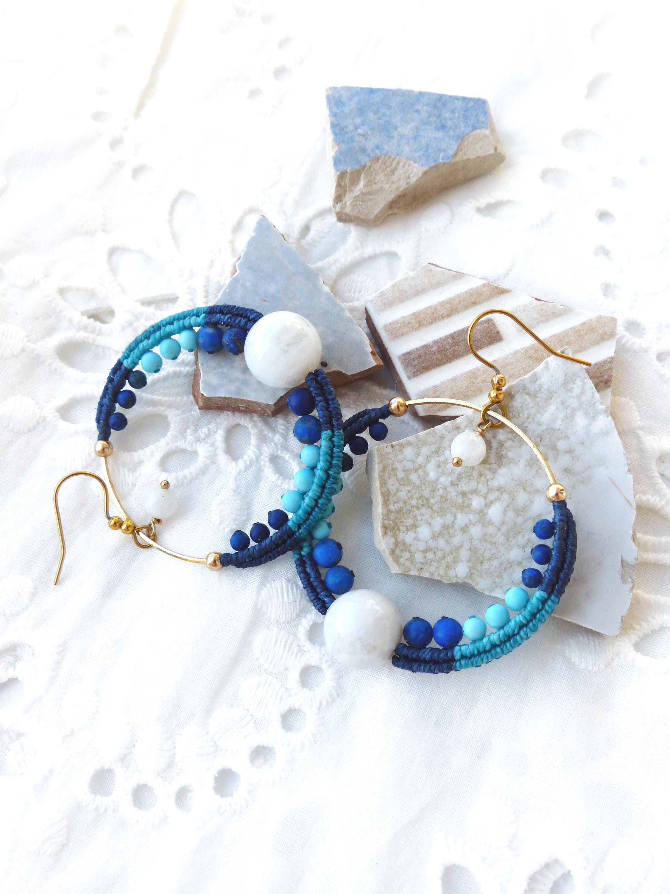 Peut inclure: Une paire de boucles d'oreilles créoles faites à la main. Les boucles d'oreilles présentent un design macramé dans des tons de bleu et turquoise, agrémenté de perles blanches et bleues. Les créoles ont des crochets d'oreilles dorés et une grosse perle blanche au centre.
