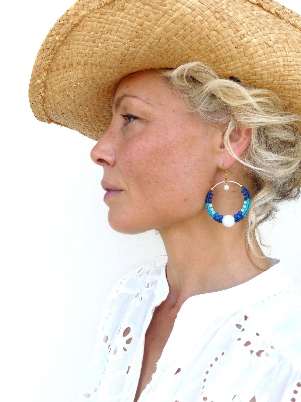Peut inclure: Une femme porte un chapeau de paille et une chemise blanche à œillets. Elle porte de grandes boucles d'oreilles créoles avec des perles bleues et blanches et une perle blanche. Les boucles d'oreilles sont attachées à un cercle en métal doré.