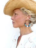 Peut inclure: Une femme porte un chapeau de paille et une chemise blanche à œillets. Elle porte de grandes boucles d'oreilles créoles avec des perles bleues et blanches et une perle blanche. Les boucles d'oreilles sont attachées à un cercle en métal doré.