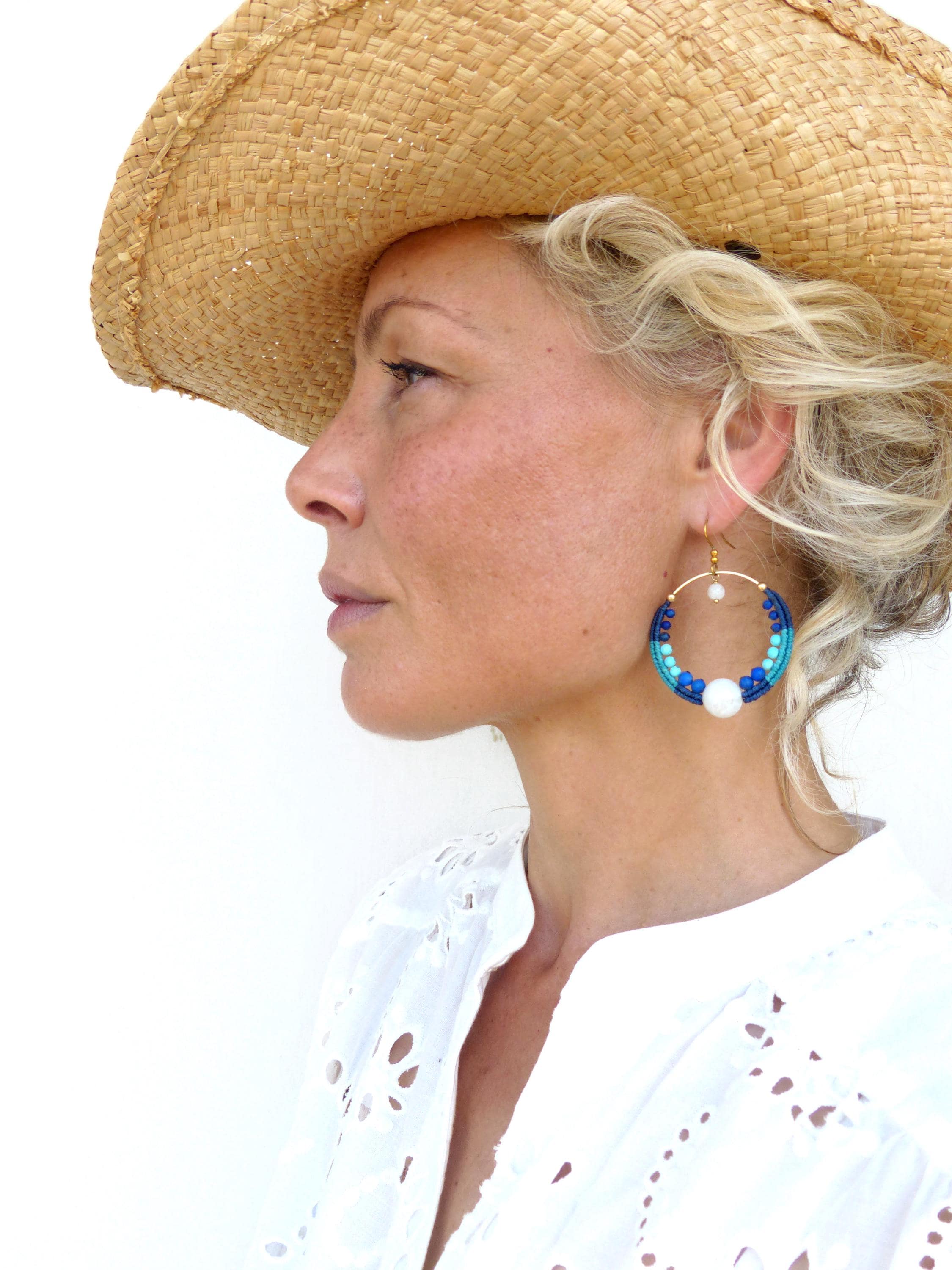 Peut inclure: Une femme porte un chapeau de paille et une chemise blanche à œillets. Elle porte de grandes boucles d'oreilles créoles avec des perles bleues et blanches et une perle blanche. Les boucles d'oreilles sont attachées à un cercle en métal doré.