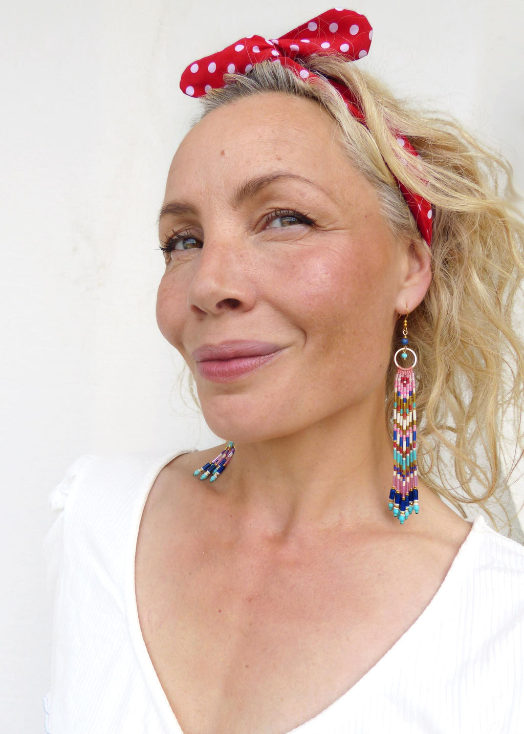 Peut inclure: Une femme porte de longues boucles d'oreilles en perles avec un motif géométrique dans les tons de rose, de bleu et de turquoise. Les boucles d'oreilles sont suspendues à des anneaux dorés. Elle porte également un bandeau à pois rouges.