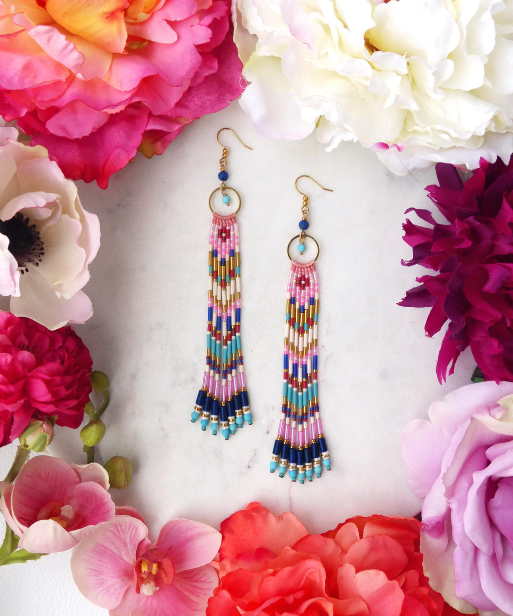 Peut inclure: Une paire de longues boucles d'oreilles perlées avec des crochets et des accents circulaires dorés. Les boucles d'oreilles présentent un motif coloré de perles roses, bleues, dorées et blanches. Le fond est orné de fleurs roses, rouges et blanches vibrantes.