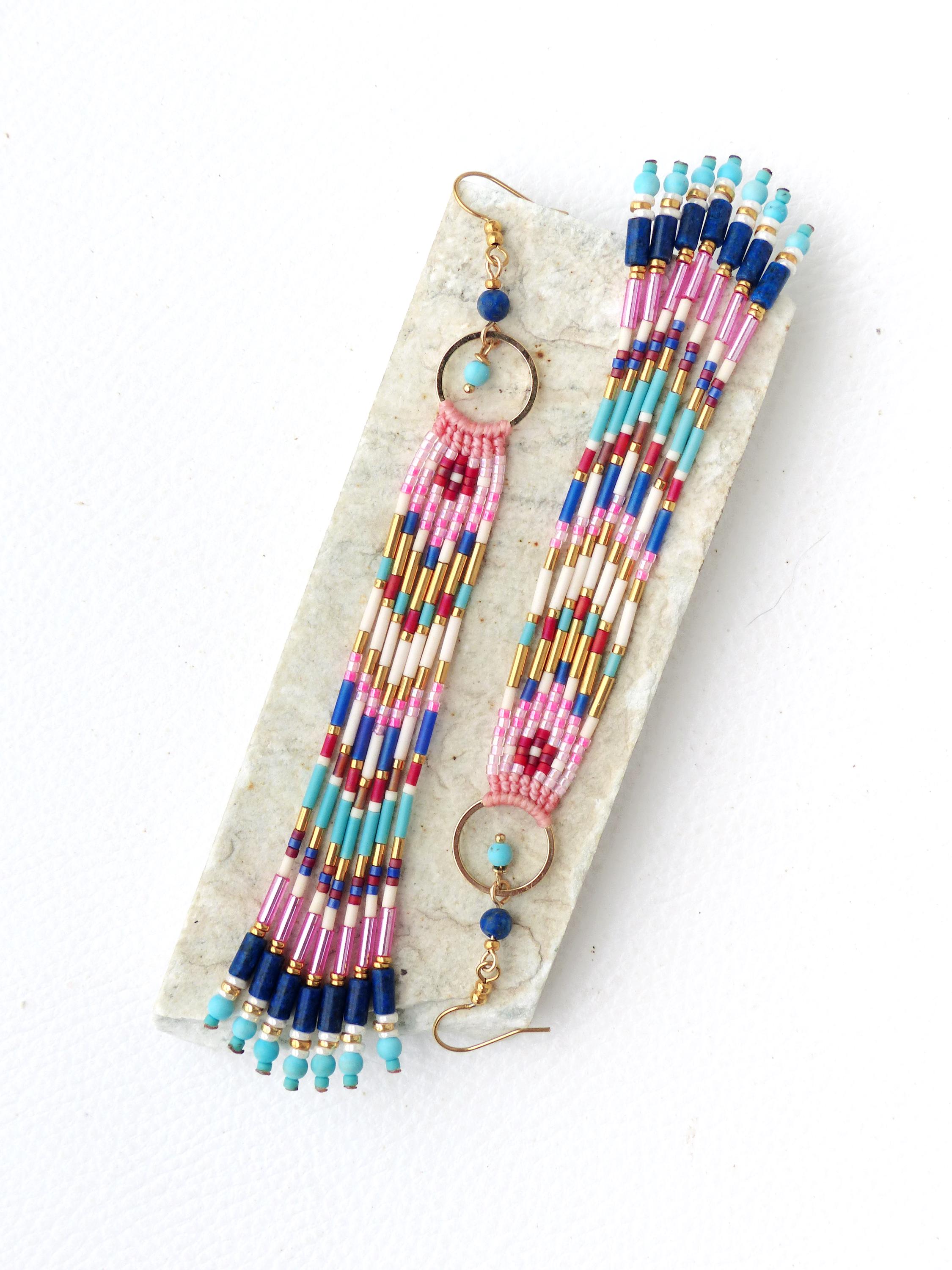 Peut inclure: Une paire de boucles d'oreilles perlées faites à la main. Les boucles d'oreilles présentent un anneau doré et une frange de perles colorées dans les tons de rose, bleu, or et blanc. Les boucles d'oreilles sont présentées sur une surface texturée de couleur claire.
