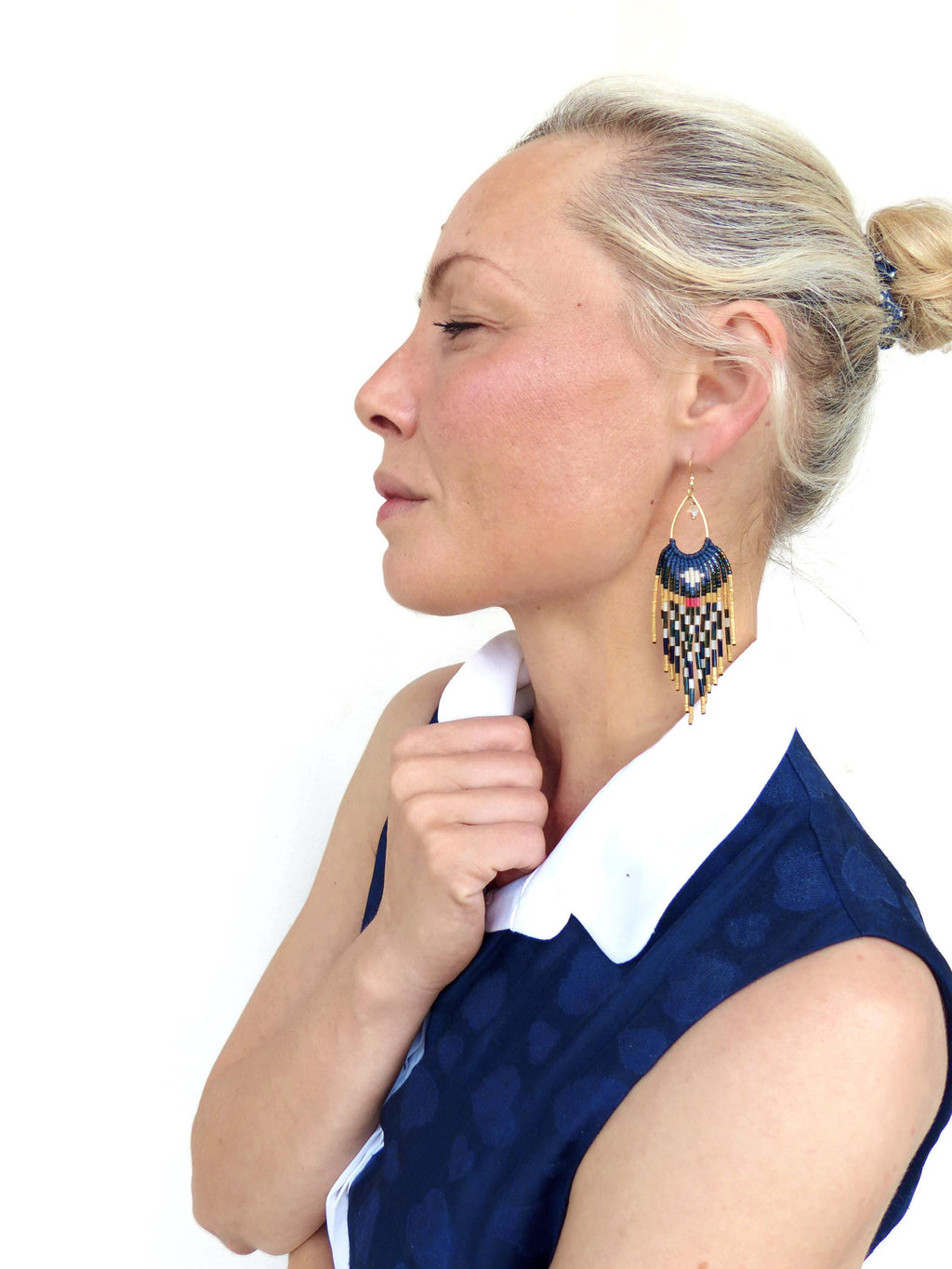 Peut inclure: Une femme porte des boucles d'oreilles en perles avec un dessus doré et un motif géométrique noir, blanc et bleu marine. La femme porte une robe bleu marine avec un col blanc.