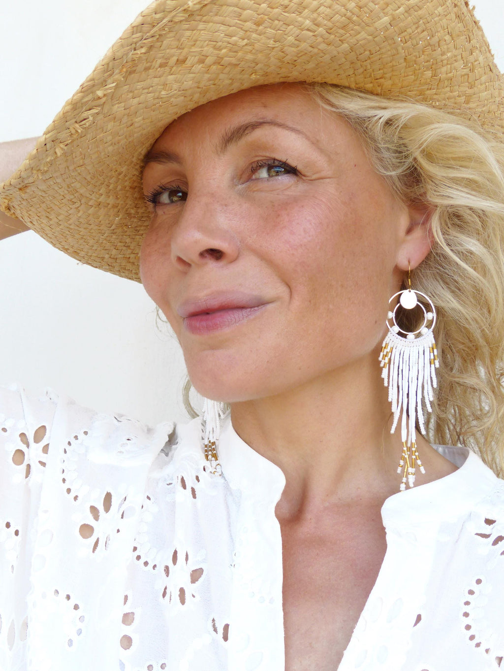 Peut inclure: Boucles d'oreilles pendantes blanches à perles avec des détails dorés. La femme porte un chapeau de paille et une chemise blanche ajourée. Les boucles d'oreilles ont un design circulaire et des perles pendantes.