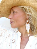 Peut inclure: Boucles d'oreilles créoles dorées avec des franges de perles blanches et dorées. Les boucles d'oreilles ont un design circulaire avec un plus petit cercle et des franges blanches en dessous. Les boucles d'oreilles sont portées avec un haut blanc à œillets et un chapeau de paille.