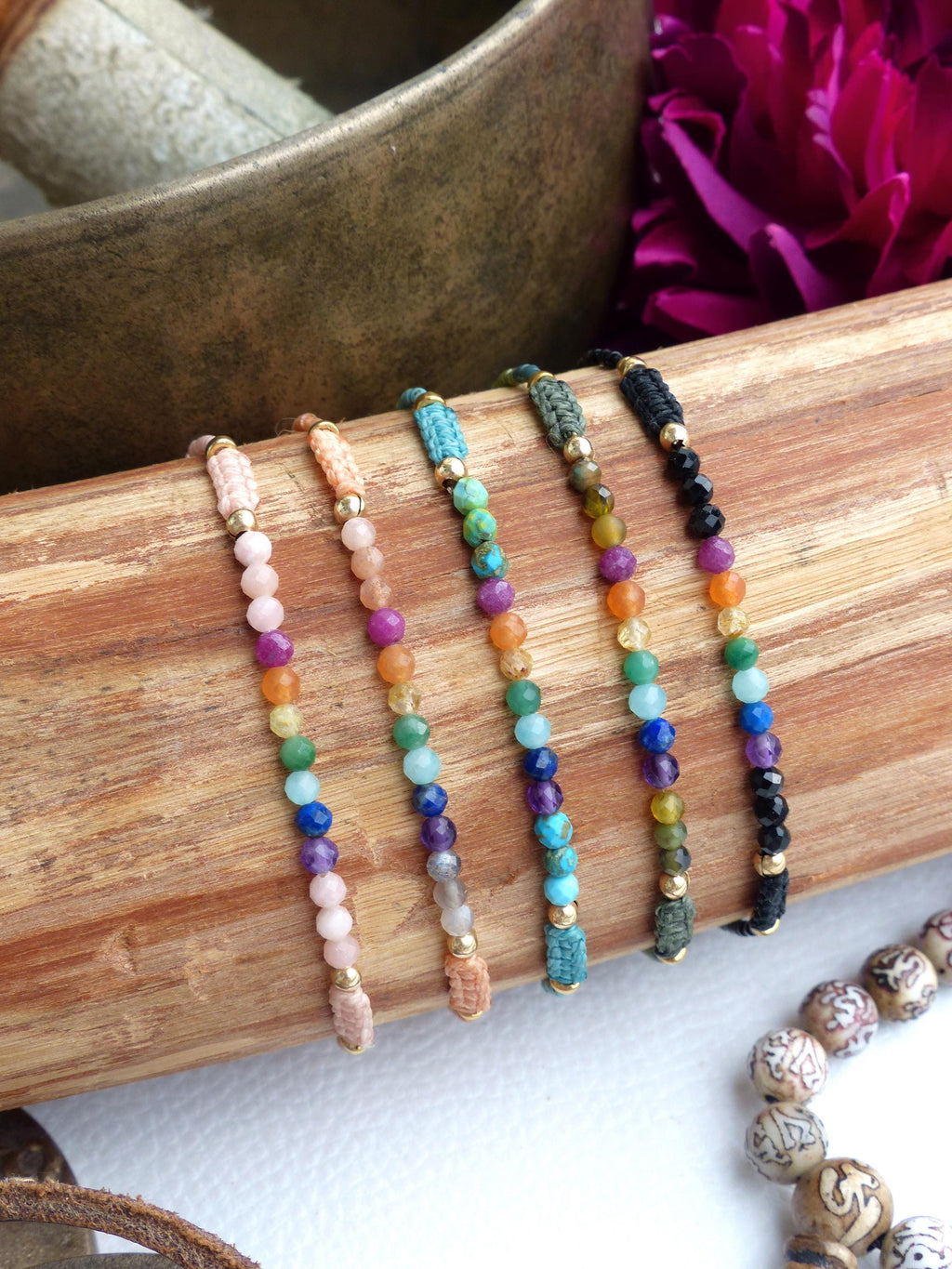 Peut inclure: Cinq bracelets de perles avec des pierres précieuses colorées et des accents dorés sont présentés sur une surface en bois. Les bracelets présentent une gamme de couleurs, dont le rose, l'orange, le vert, le bleu et le violet. Les bracelets sont enfilés avec un cordon.