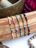 Peut inclure: Cinq bracelets de perles avec des pierres précieuses colorées et des accents dorés sont présentés sur une surface en bois. Les bracelets présentent une gamme de couleurs, dont le rose, l'orange, le vert, le bleu et le violet. Les bracelets sont enfilés avec un cordon.