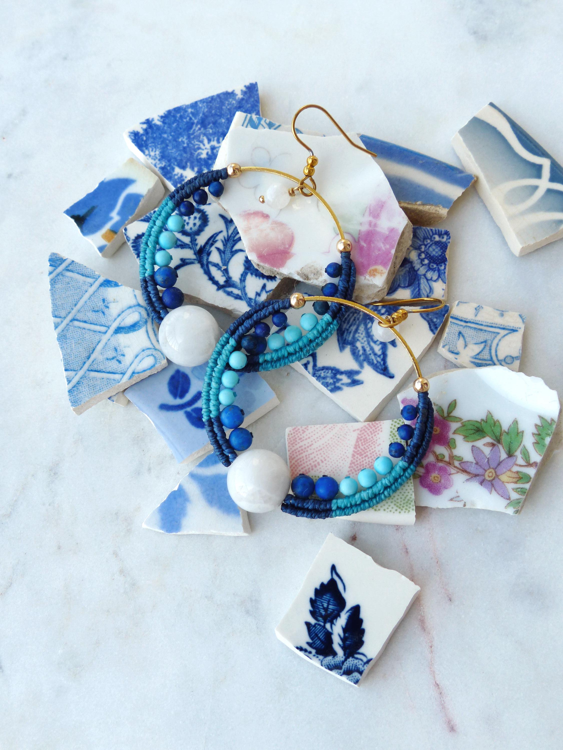 Peut inclure: Boucles d'oreilles créoles faites à la main avec un design macramé dans des tons de bleu et turquoise, ornées de perles blanches. Les boucles d'oreilles reposent sur des morceaux de céramique bleus et blancs cassés.