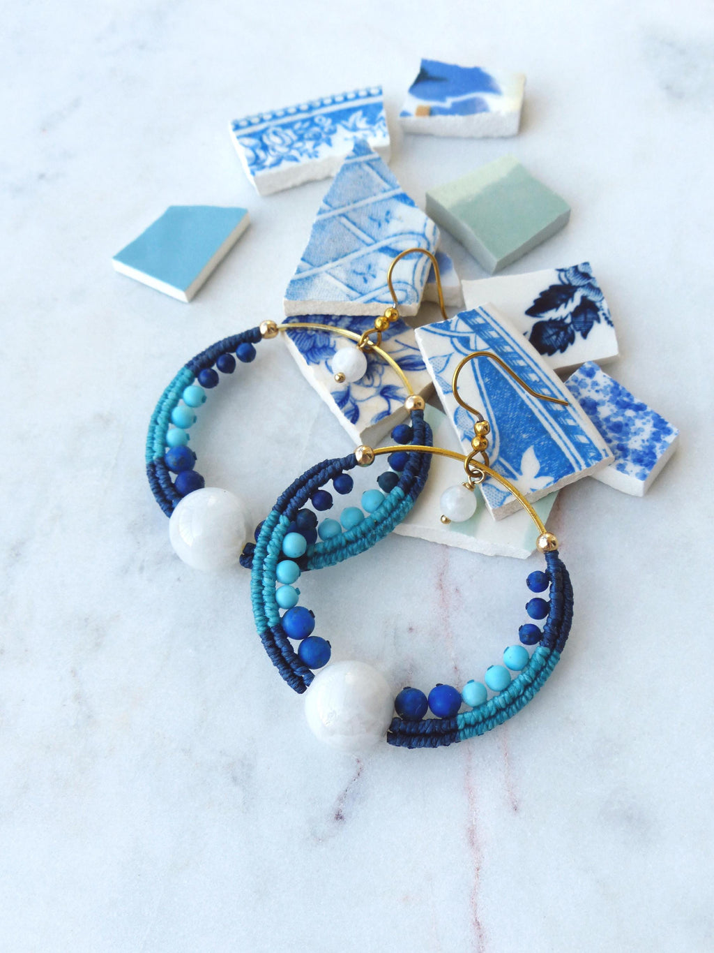 Peut inclure: Boucles d'oreilles créoles faites à la main avec un design macramé dans les tons de bleu et blanc. Les boucles d'oreilles comportent une grosse perle blanche au centre et de petites perles bleues. Des crochets d'oreilles dorés complètent le design. Des éclats de céramique bleus et blancs décoratifs sont en arrière-plan.