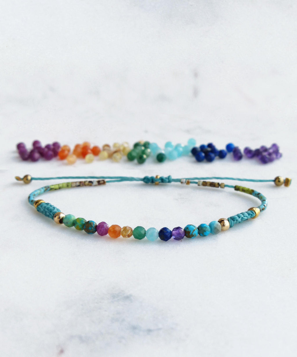 Peut inclure: Un bracelet fait à la main avec une rangée de perles colorées dans un spectre arc-en-ciel. Le bracelet est monté sur un cordon turquoise avec des accents dorés. Des perles supplémentaires sont disposées au-dessus.
