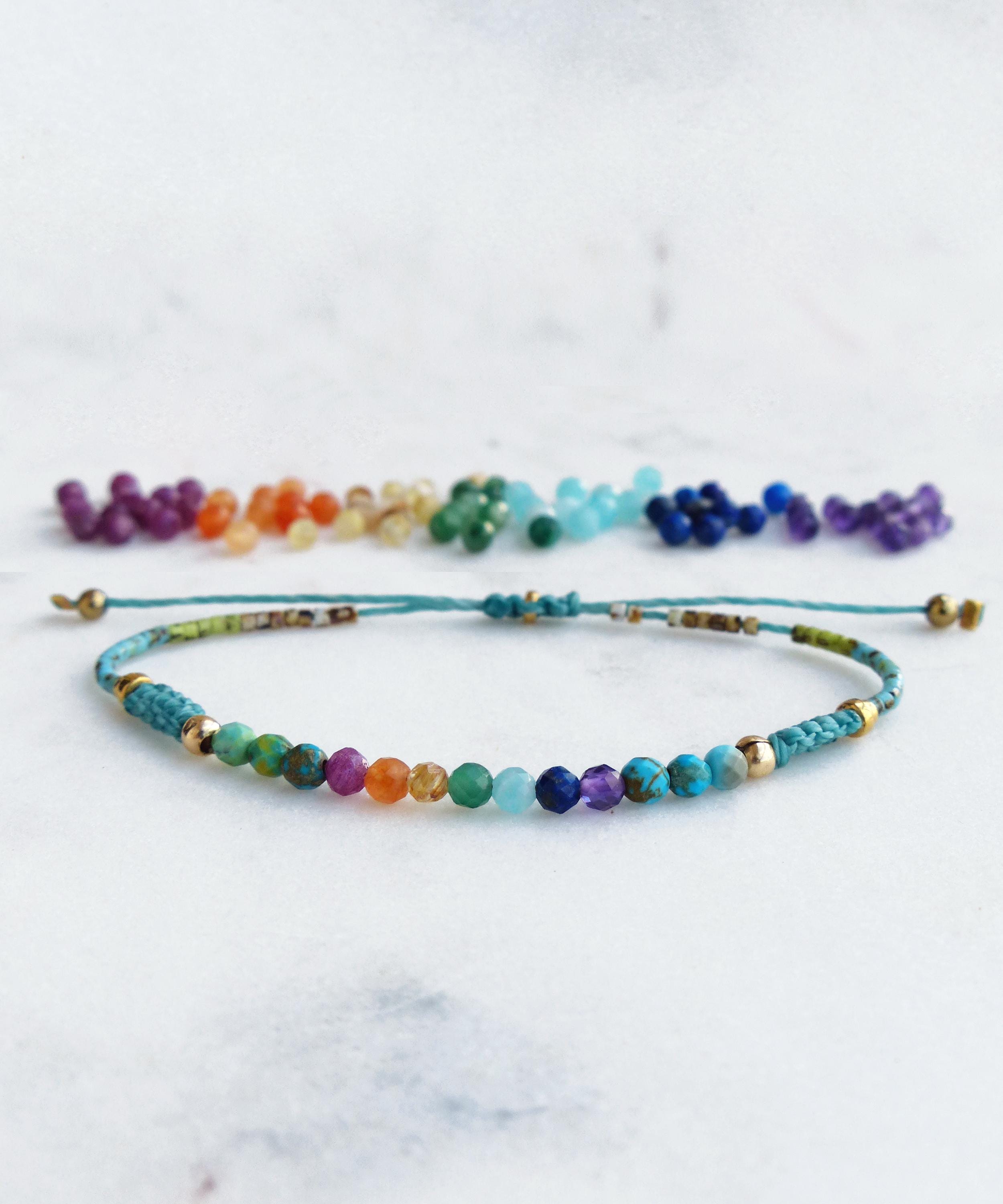 Peut inclure: Un bracelet fait à la main avec une rangée de perles colorées dans un spectre arc-en-ciel. Le bracelet est monté sur un cordon turquoise avec des accents dorés. Des perles supplémentaires sont disposées au-dessus.