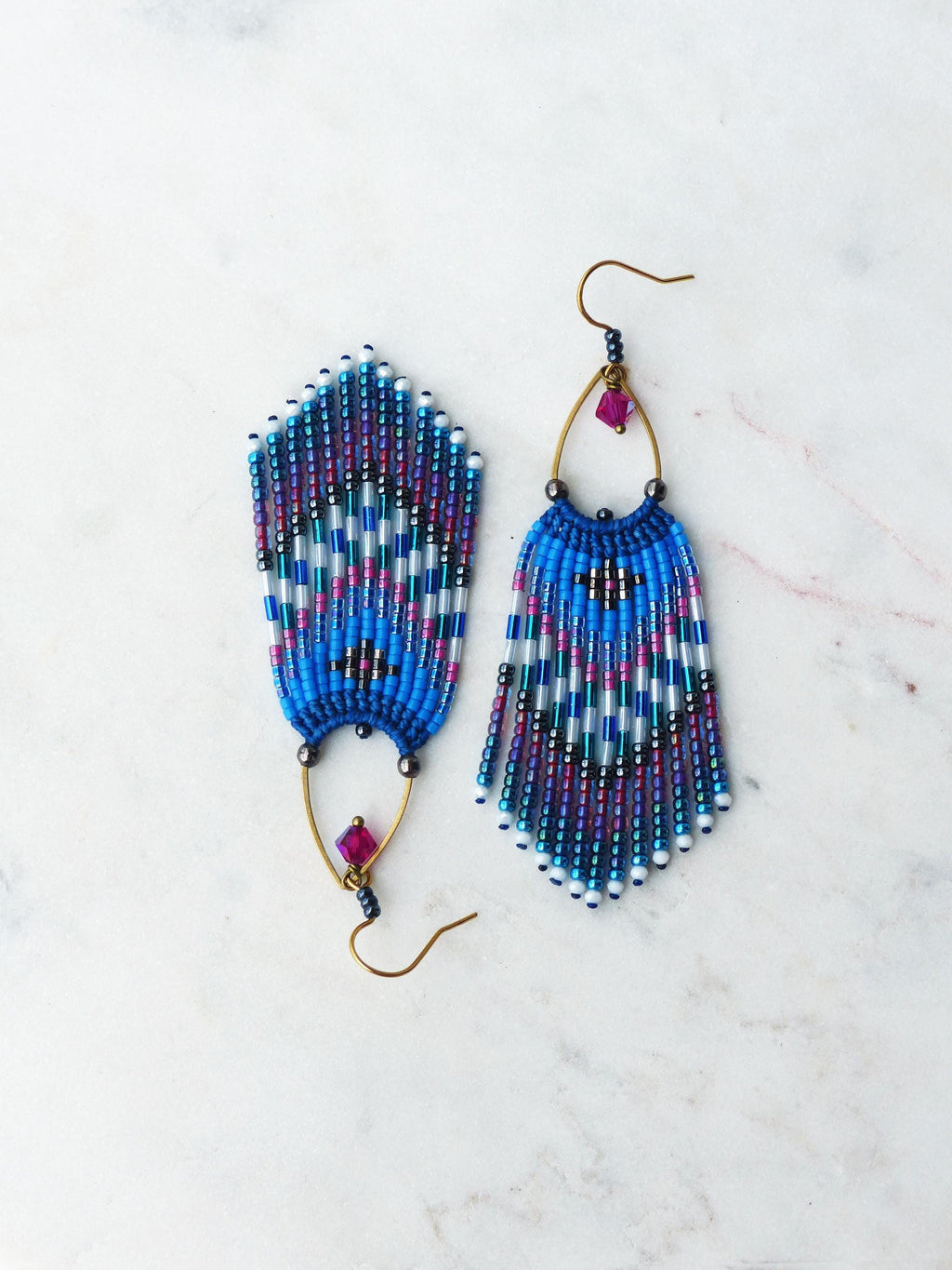 Peut inclure: Paire de boucles d'oreilles en perles faites à la main. Elles présentent un motif bleu, blanc et violet avec un design à franges. Chaque boucle d'oreille a un crochet doré et une petite perle rose carrée.
