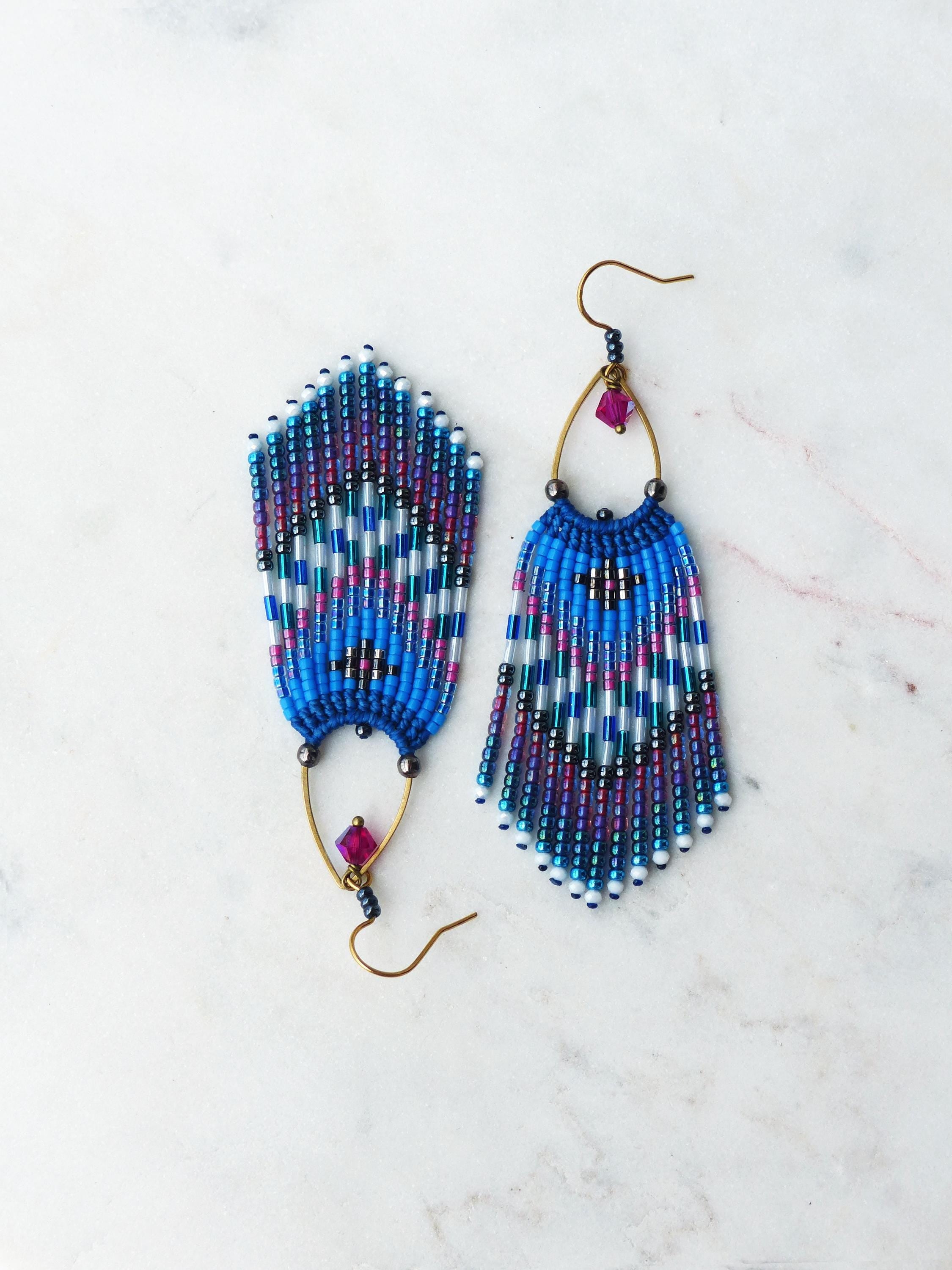 Peut inclure: Paire de boucles d'oreilles en perles faites à la main. Elles présentent un motif bleu, blanc et violet avec un design à franges. Chaque boucle d'oreille a un crochet doré et une petite perle rose carrée.