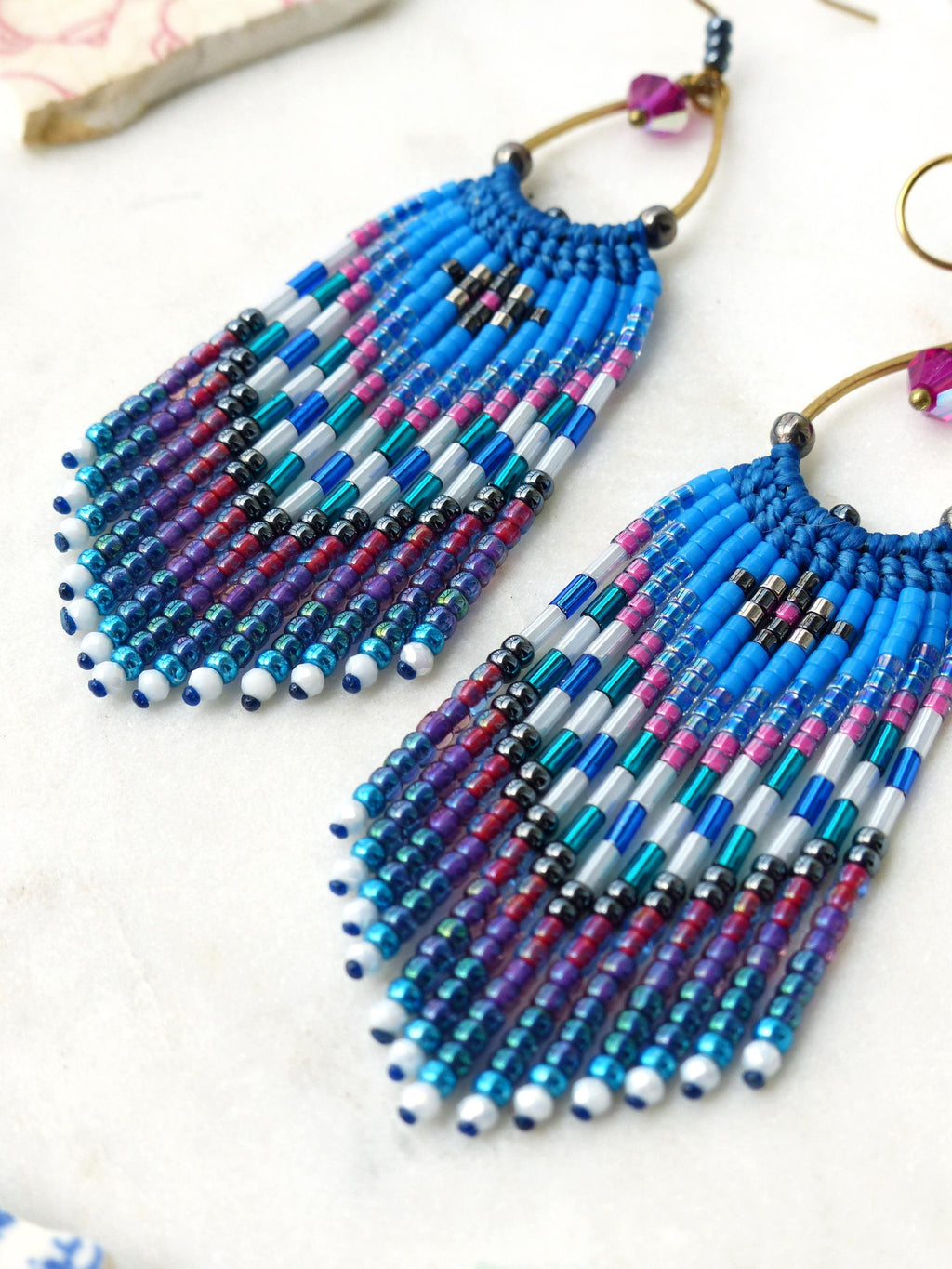 Peut inclure: Une paire de boucles d'oreilles en perles aux tons bleus, violets et roses. Les boucles d'oreilles présentent un design à franges avec un dessus tissé et des cerceaux dorés. Les perles sont disposées en rangées verticales, créant un effet texturé.
