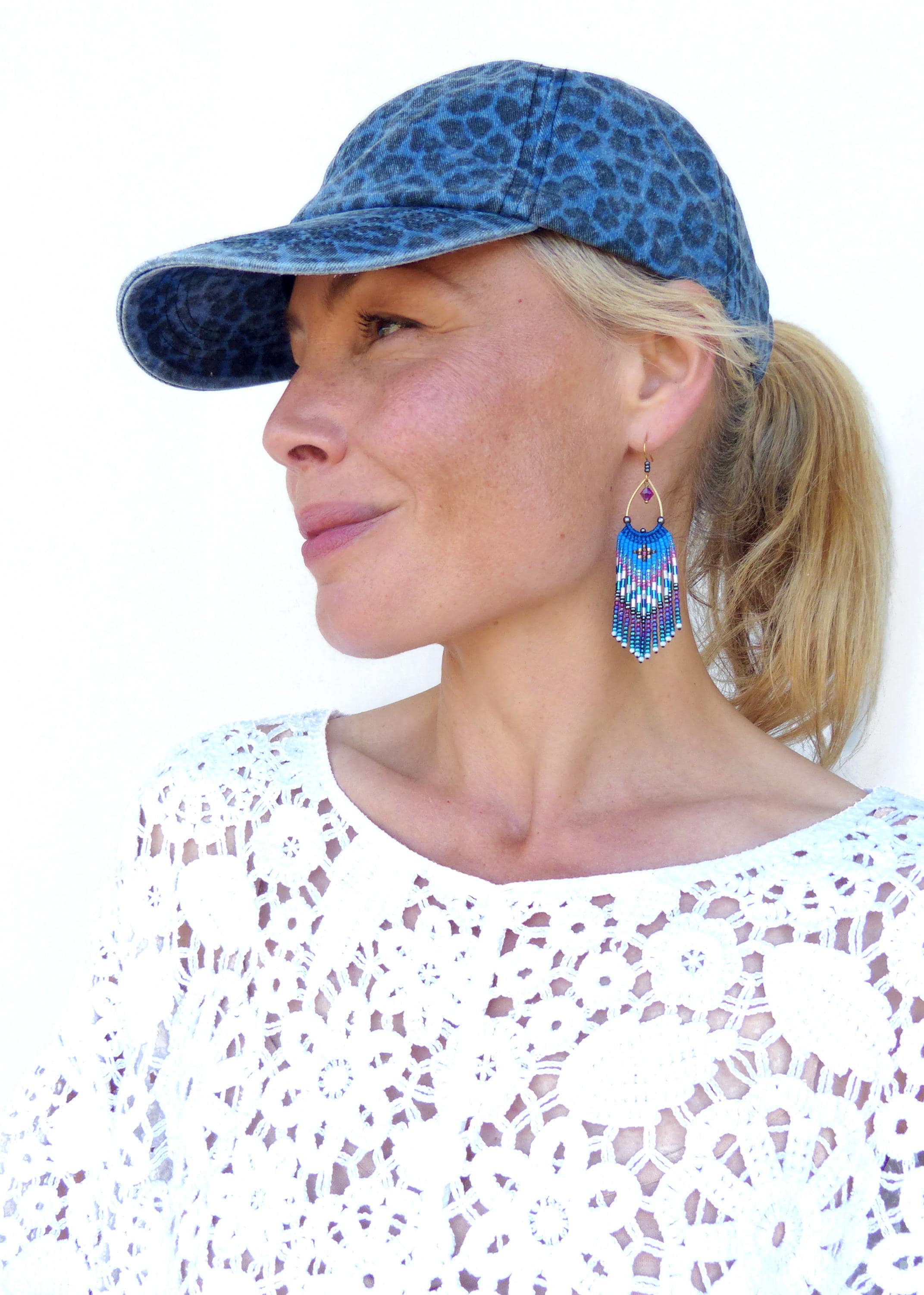 Peut inclure: Casquette de baseball en jean bleu avec un motif léopard. La photo présente des boucles d'oreilles en perles avec des franges bleues, violettes et blanches. Les boucles d'oreilles ont un cerceau doré et une petite perle violette. La femme porte un haut en dentelle blanche.
