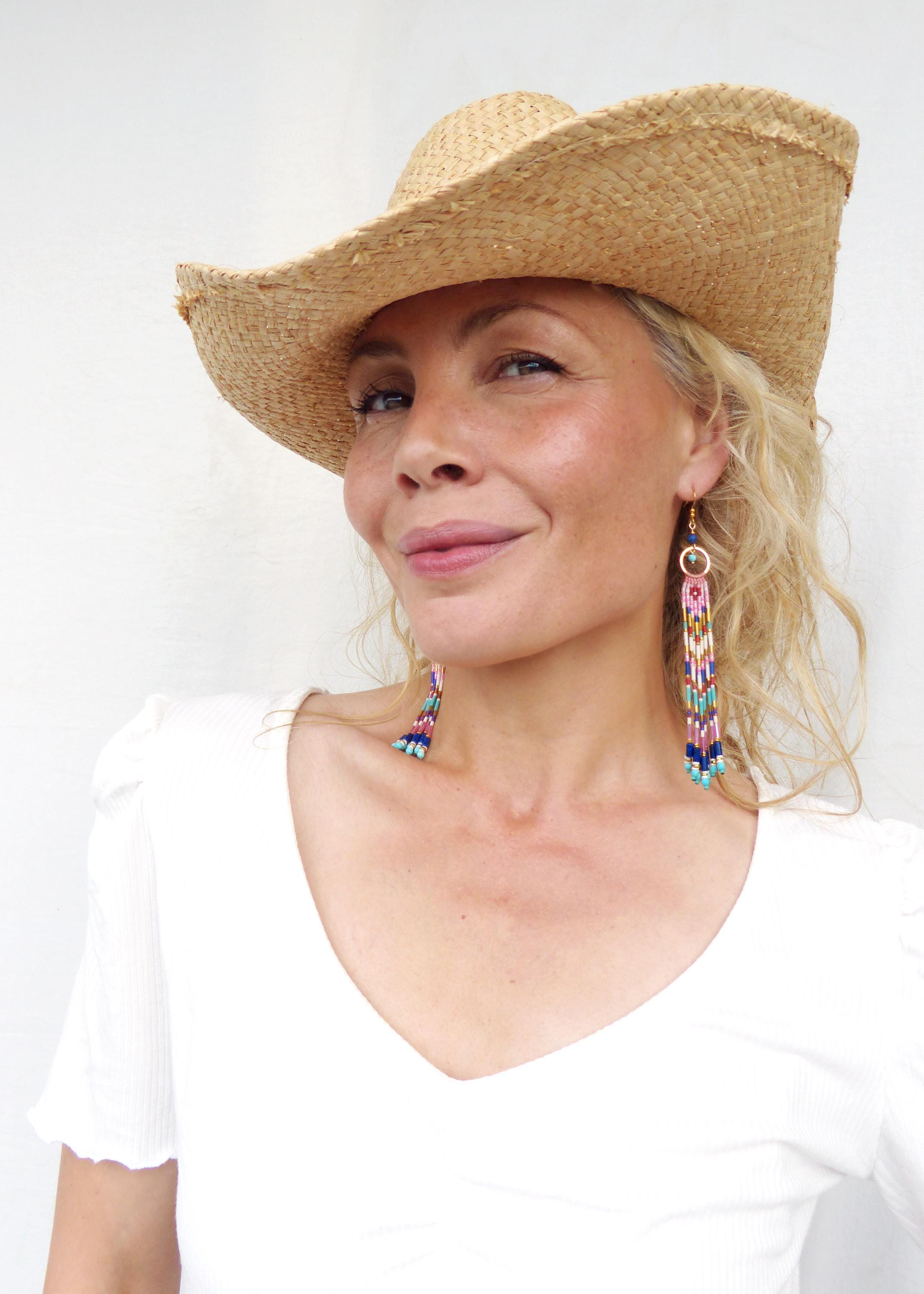 Peut inclure: Une femme porte un chapeau de paille et de longues boucles d'oreilles perlées. Les boucles d'oreilles présentent un mélange de perles roses, bleues et jaunes. Elle porte un haut blanc avec un col en V.