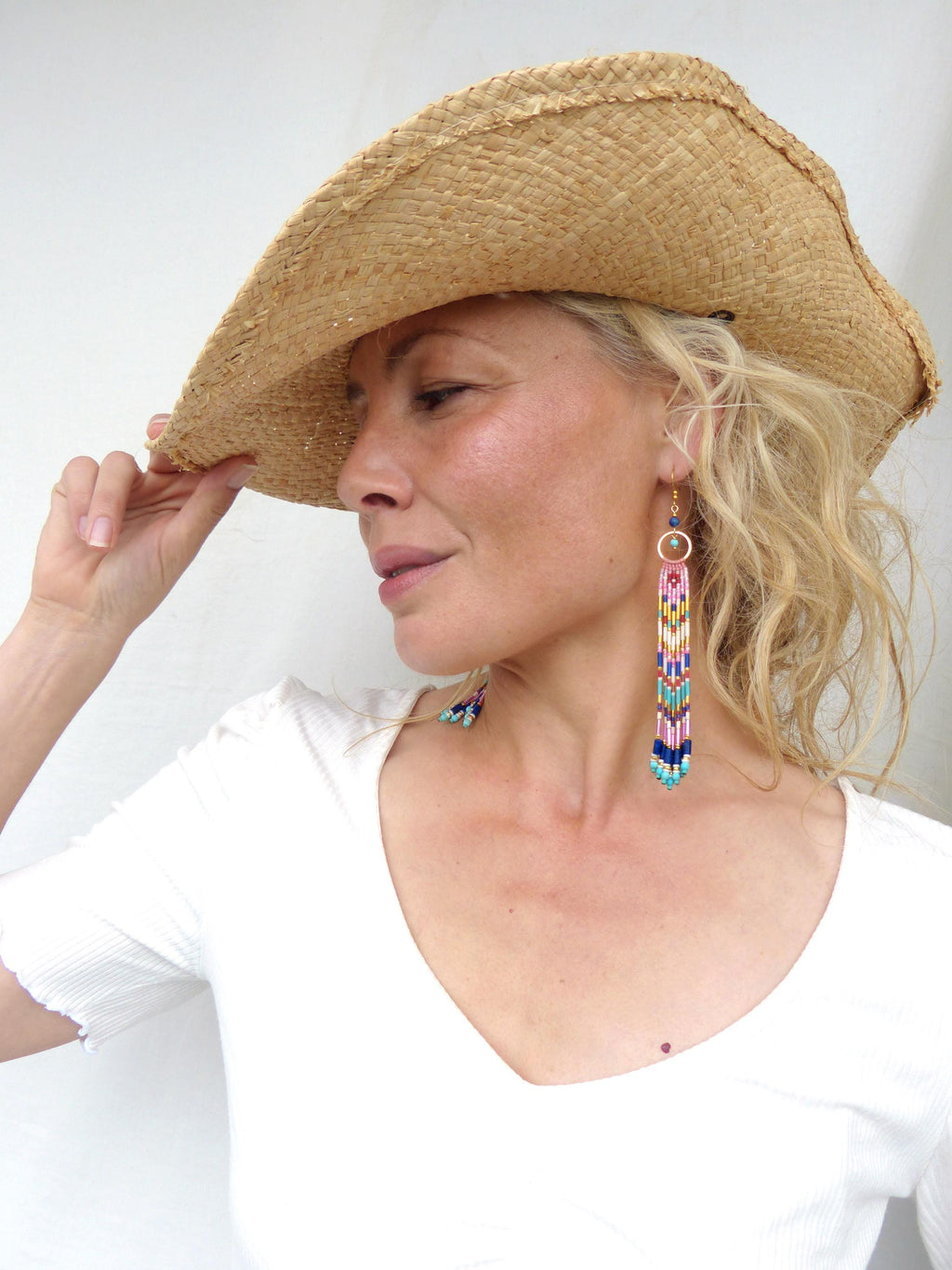 Peut inclure: Une femme porte un chapeau de paille à larges bords et de longues boucles d'oreilles perlées. Les boucles d'oreilles présentent un cerceau doré et un motif vertical avec des perles roses, bleues et jaunes. Elle porte un haut blanc à col en V.
