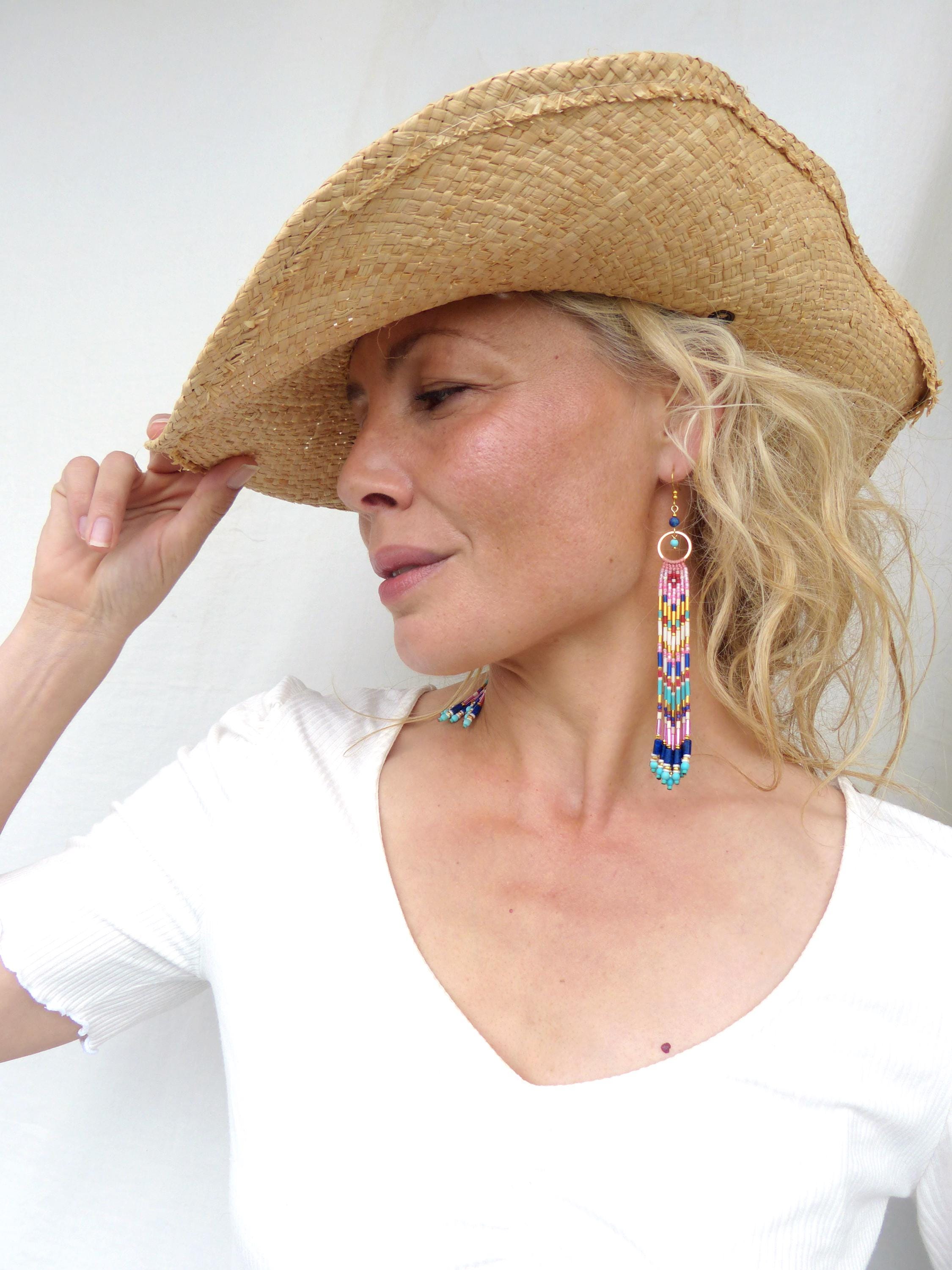 Peut inclure: Une femme porte un chapeau de paille à larges bords et de longues boucles d'oreilles perlées. Les boucles d'oreilles présentent un cerceau doré et un motif vertical avec des perles roses, bleues et jaunes. Elle porte un haut blanc à col en V.