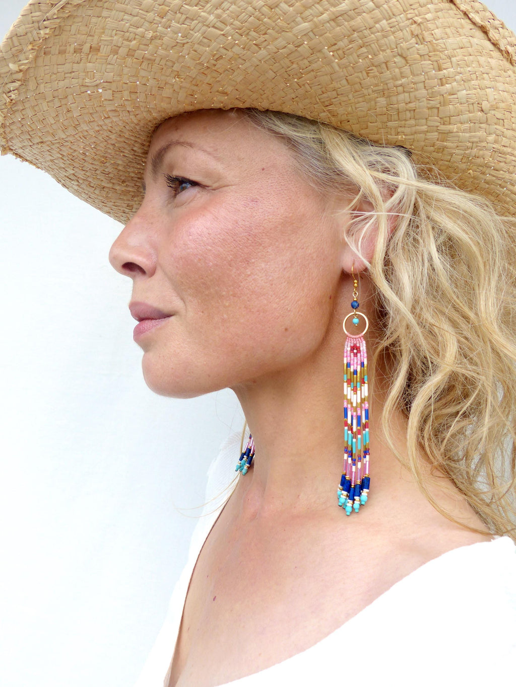Peut inclure: Portrait de profil d'une personne portant un chapeau de paille et de longues boucles d'oreilles perlées. Les boucles d'oreilles sont composées d'un cercle doré avec une pierre bleue et d'une longue frange de perles colorées.