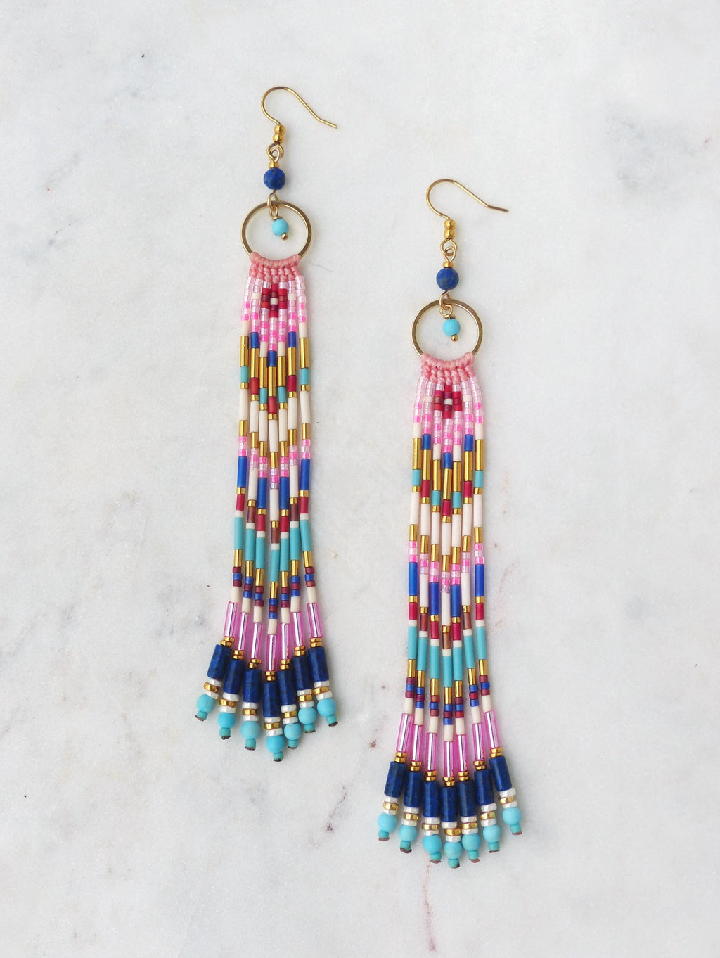 Peut inclure: Une paire de boucles d'oreilles en perles avec un crochet doré et un accent doré circulaire. Les boucles d'oreilles présentent un motif de perles roses, blanches, bleues et dorées. Le bas des boucles d'oreilles a une frange de perles bleu foncé et turquoise.