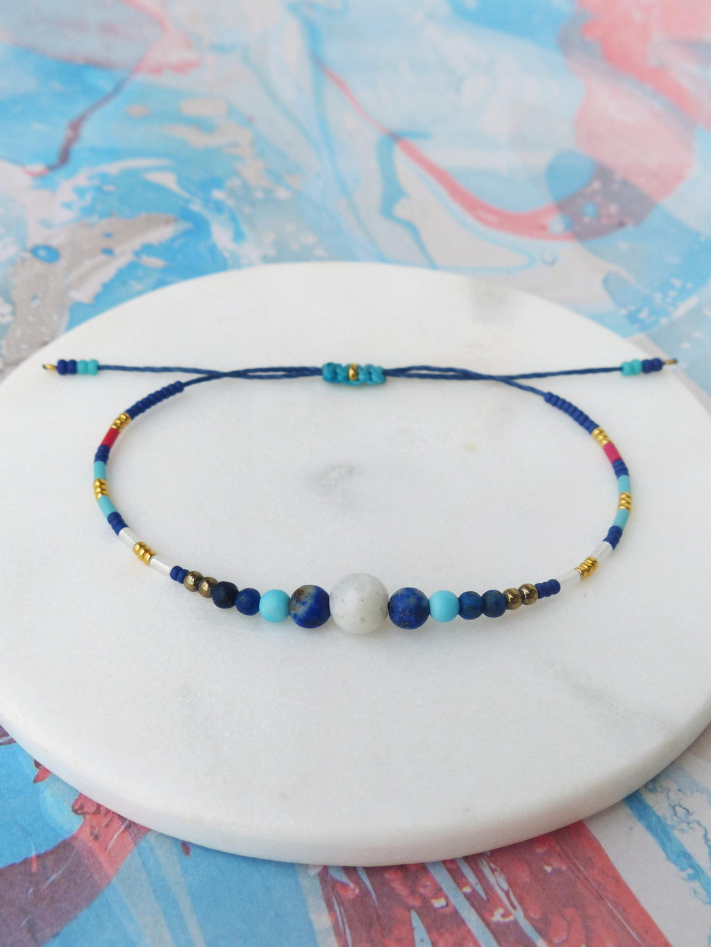 Peut inclure: Bracelet réglable avec un cordon bleu foncé orné d'une série de perles colorées. Le bracelet comprend des perles bleues, blanches, rouges et dorées, avec une grosse perle blanche centrale et de petites perles bleues.
