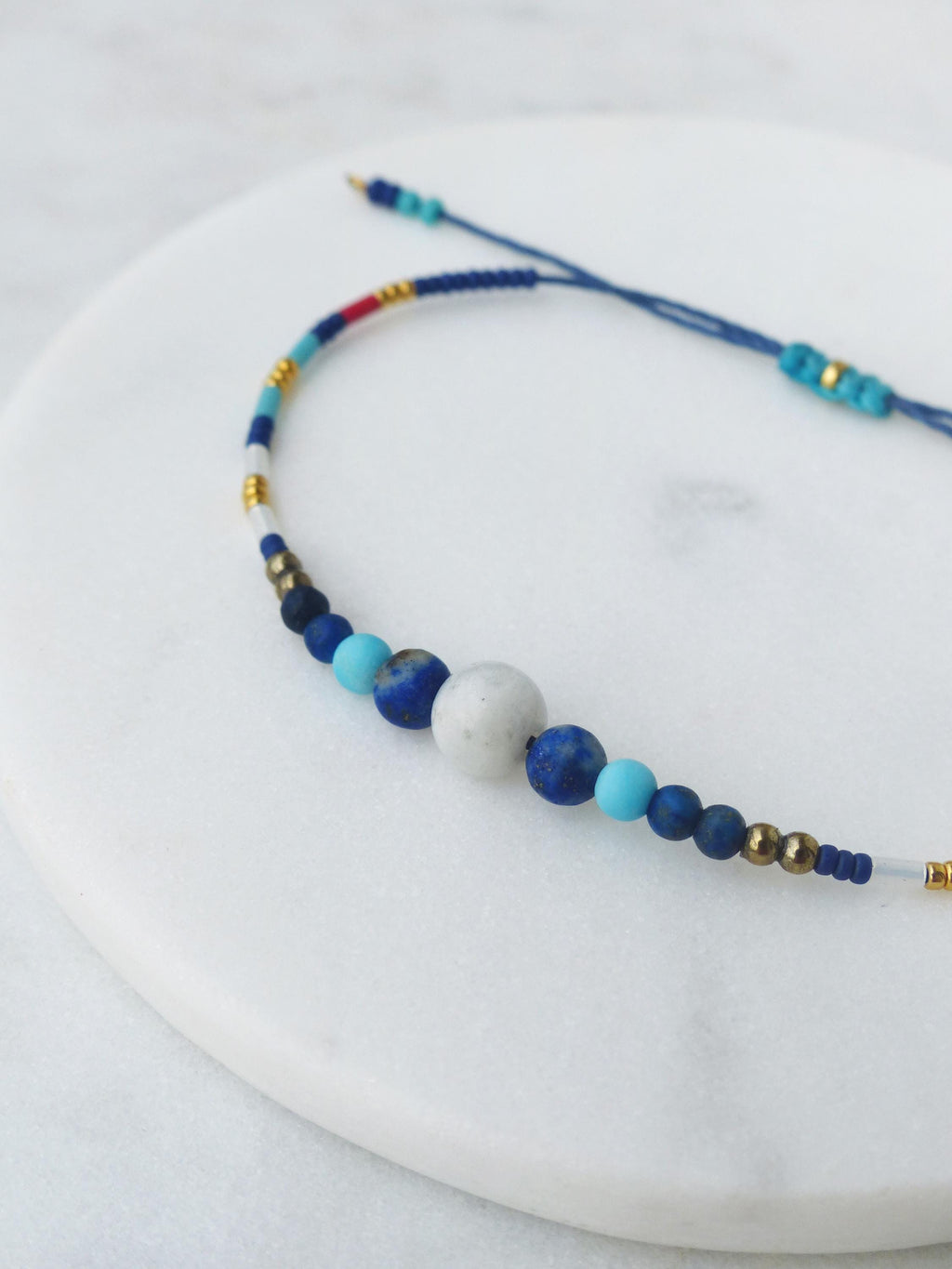 Peut inclure: Un bracelet de perles bleues et blanches avec une fermeture coulissante. Le bracelet présente une variété de perles dans les tons de bleu, blanc et or. Le bracelet est présenté sur une surface blanche.