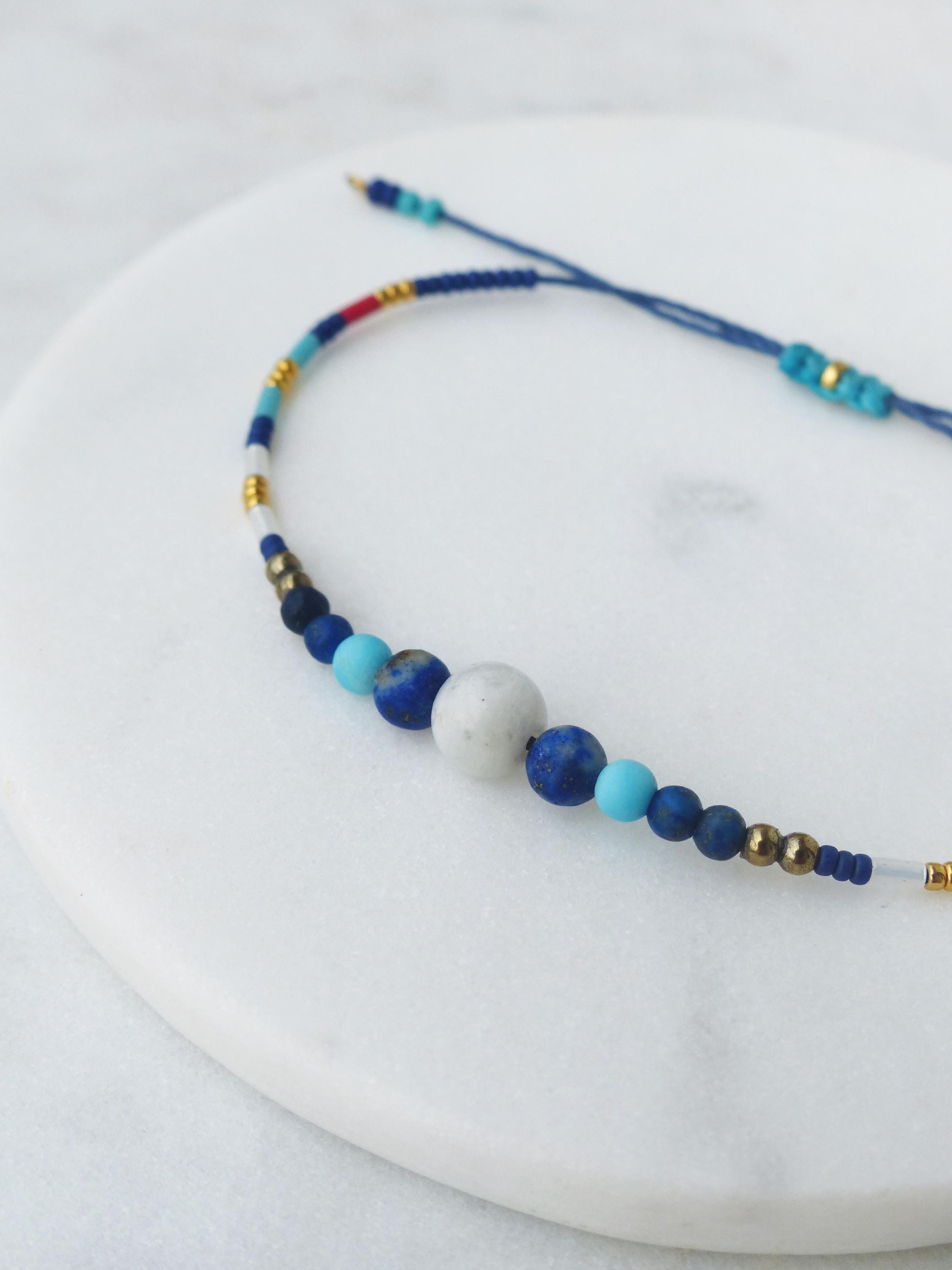 Peut inclure: Un bracelet de perles bleues et blanches avec une fermeture coulissante. Le bracelet présente une variété de perles dans les tons de bleu, blanc et or. Le bracelet est présenté sur une surface blanche.