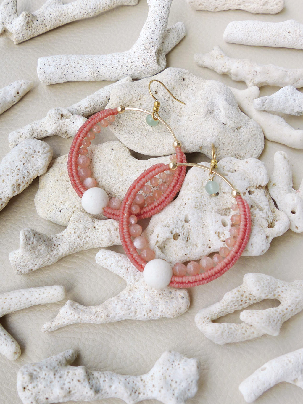 Peut inclure: Boucles d'oreilles créoles roses perlées avec crochets et accents dorés. Les créoles présentent un design tissé avec de petites perles roses et de plus grosses perles blanches. De petites perles vertes sont également présentes. Les boucles d'oreilles sont présentées sur du corail blanc.
