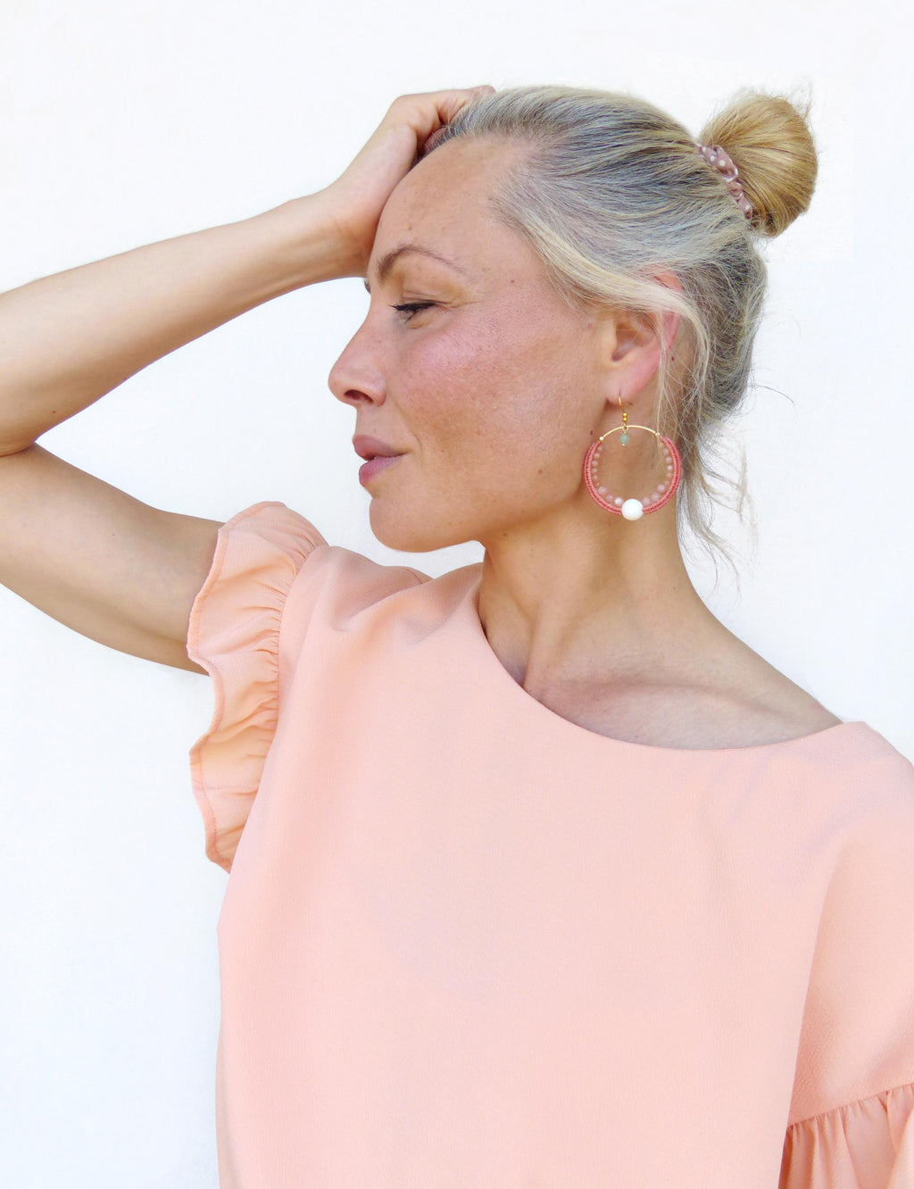 Peut inclure: Une femme porte de grandes boucles d'oreilles roses avec un cadre doré et un accent de perle blanche. Les boucles d'oreilles sont au centre de l'attention, complétées par un haut pêche à manches volantées. Les cheveux de la femme sont coiffés en chignon.