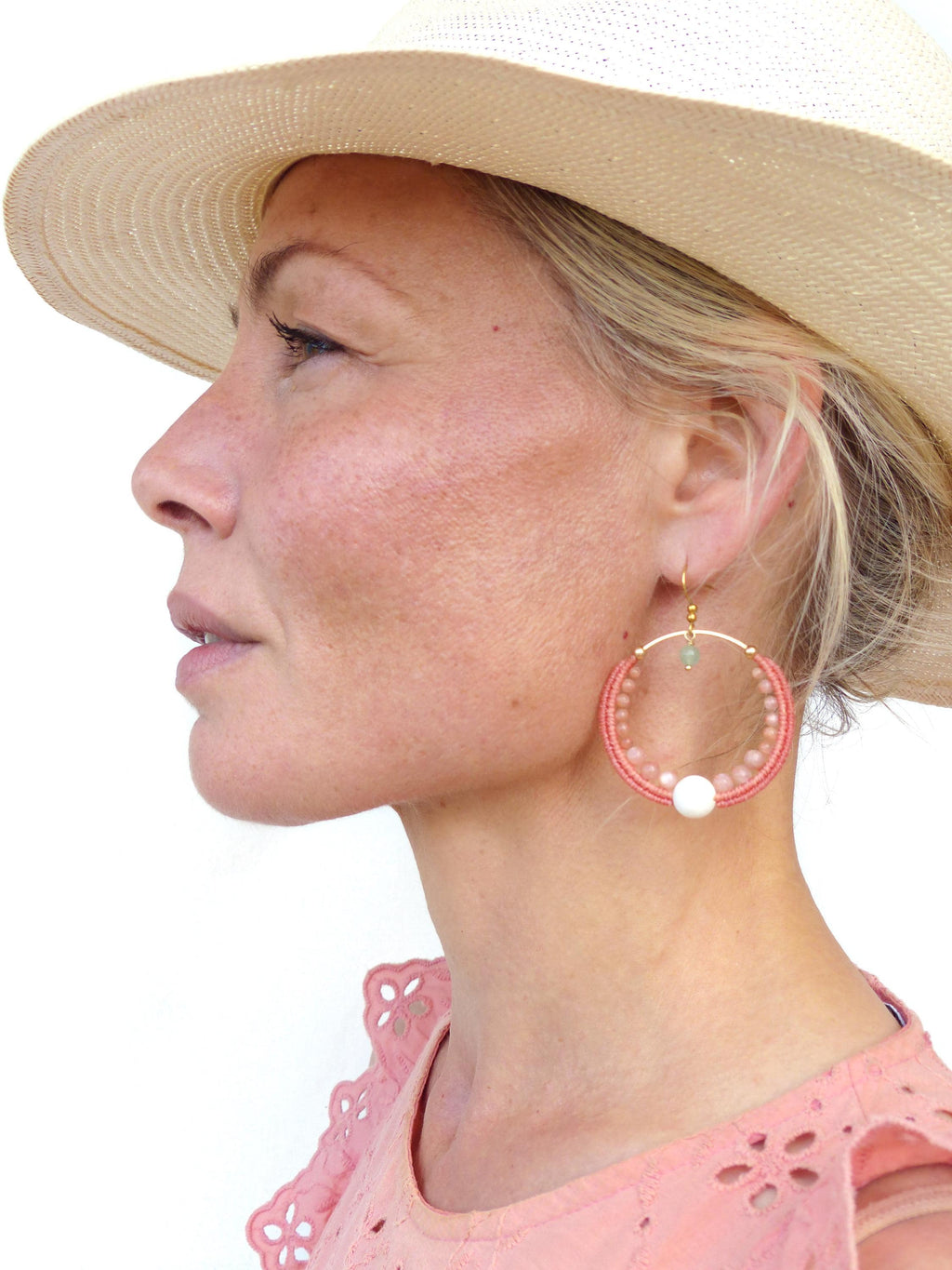 Peut inclure: Boucles d'oreilles créoles dorées avec perles corail et une grosse perle blanche. Les boucles d'oreilles ont une petite perle turquoise et sont portées avec un haut rose et un chapeau de paille.