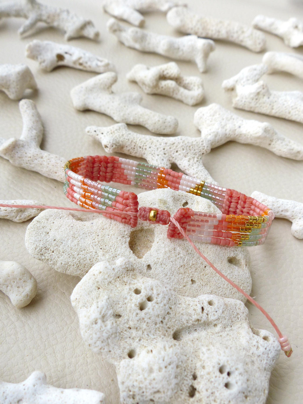 Peut inclure: Un bracelet de perles colorées aux tons rose, orange et blanc, avec des accents dorés. Le bracelet est présenté sur une surface blanche avec des décorations en forme de corail. Le bracelet est noué avec une ficelle rose.