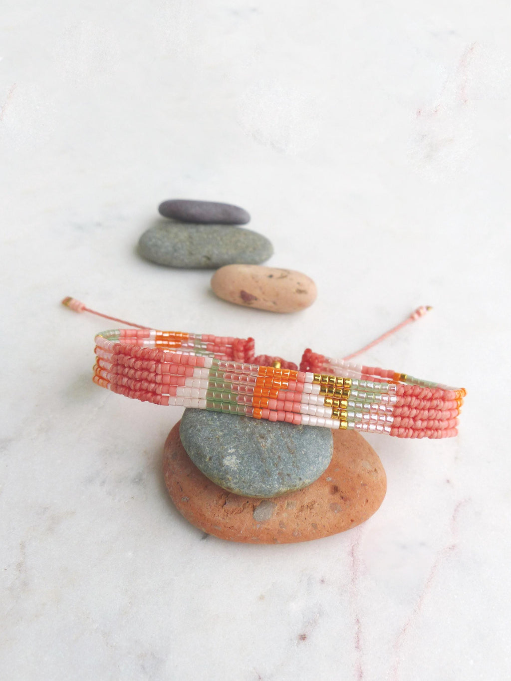 Peut inclure: Un bracelet de perles aux tons rose, orange, blanc et vert, avec des détails dorés. Le bracelet est présenté sur une pile de pierres lisses sur fond blanc. Le bracelet est de conception tissée.