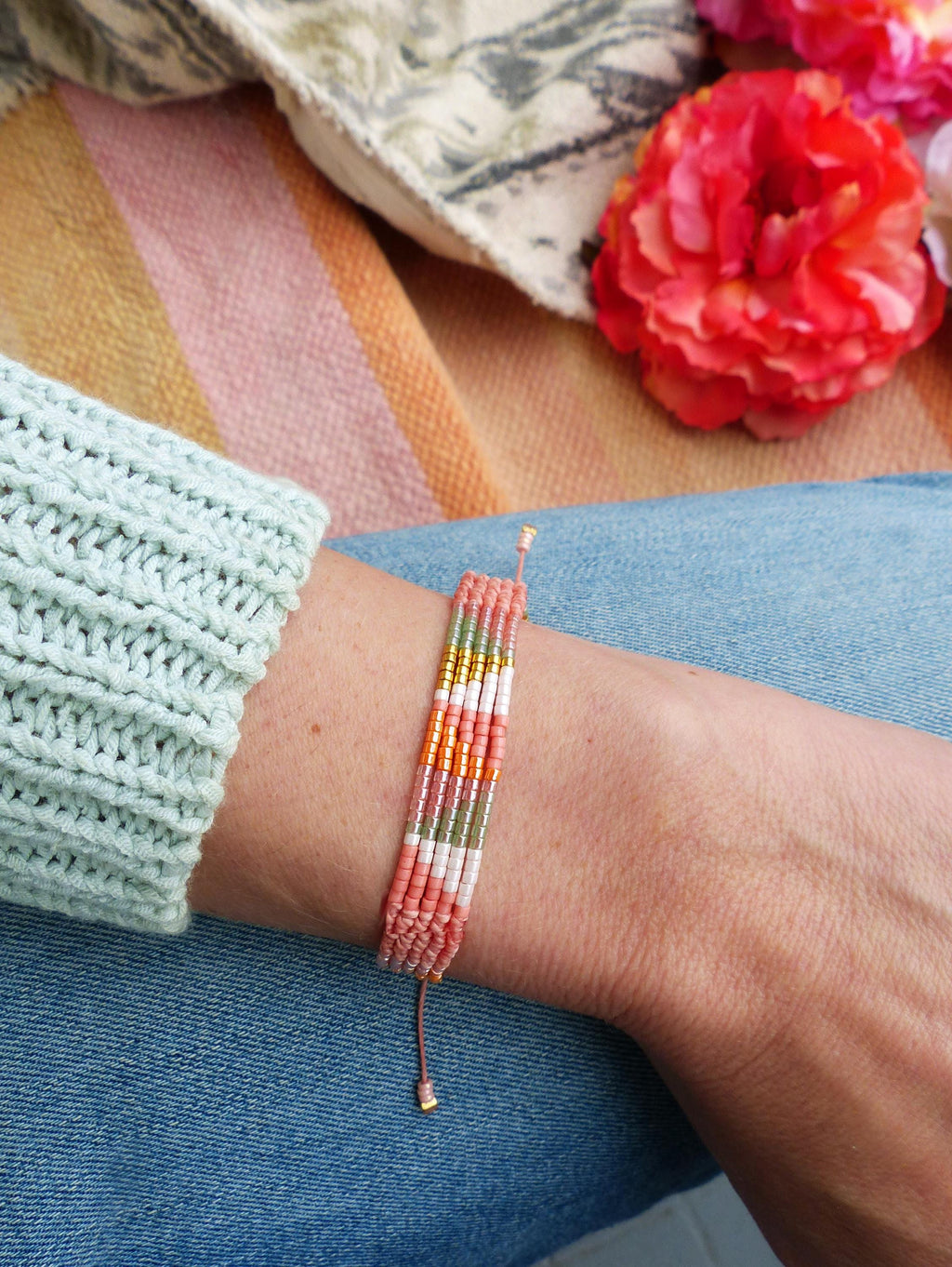 Peut inclure: Un bracelet de perles multicolores, avec des perles roses, dorées, vertes et blanches. Le bracelet est porté sur un poignet, avec une manche de pull bleu clair. Le fond présente un tissu rayé et des fleurs rouges.