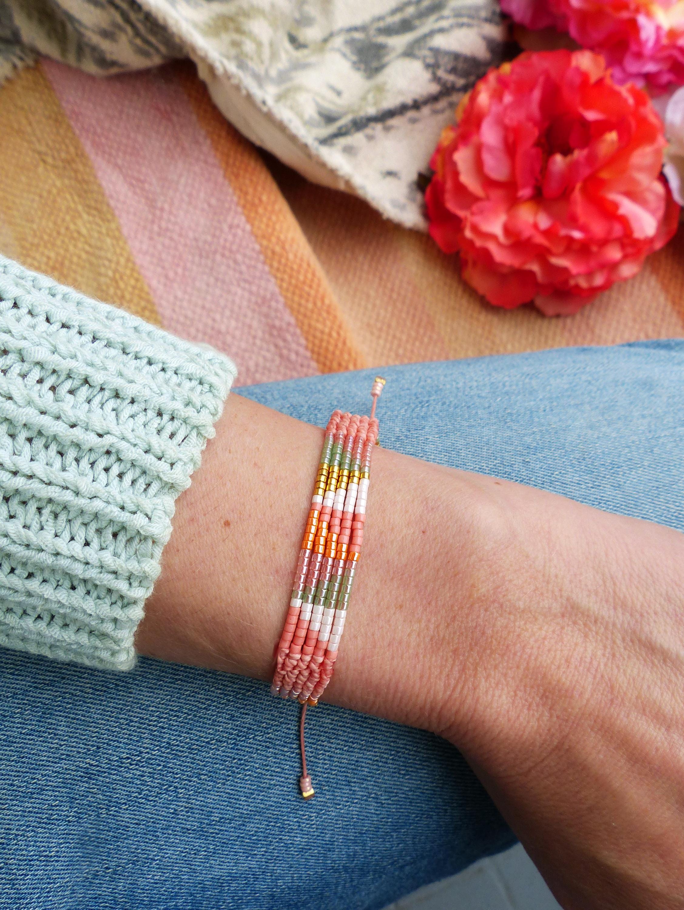Peut inclure: Un bracelet de perles multicolores, avec des perles roses, dorées, vertes et blanches. Le bracelet est porté sur un poignet, avec une manche de pull bleu clair. Le fond présente un tissu rayé et des fleurs rouges.