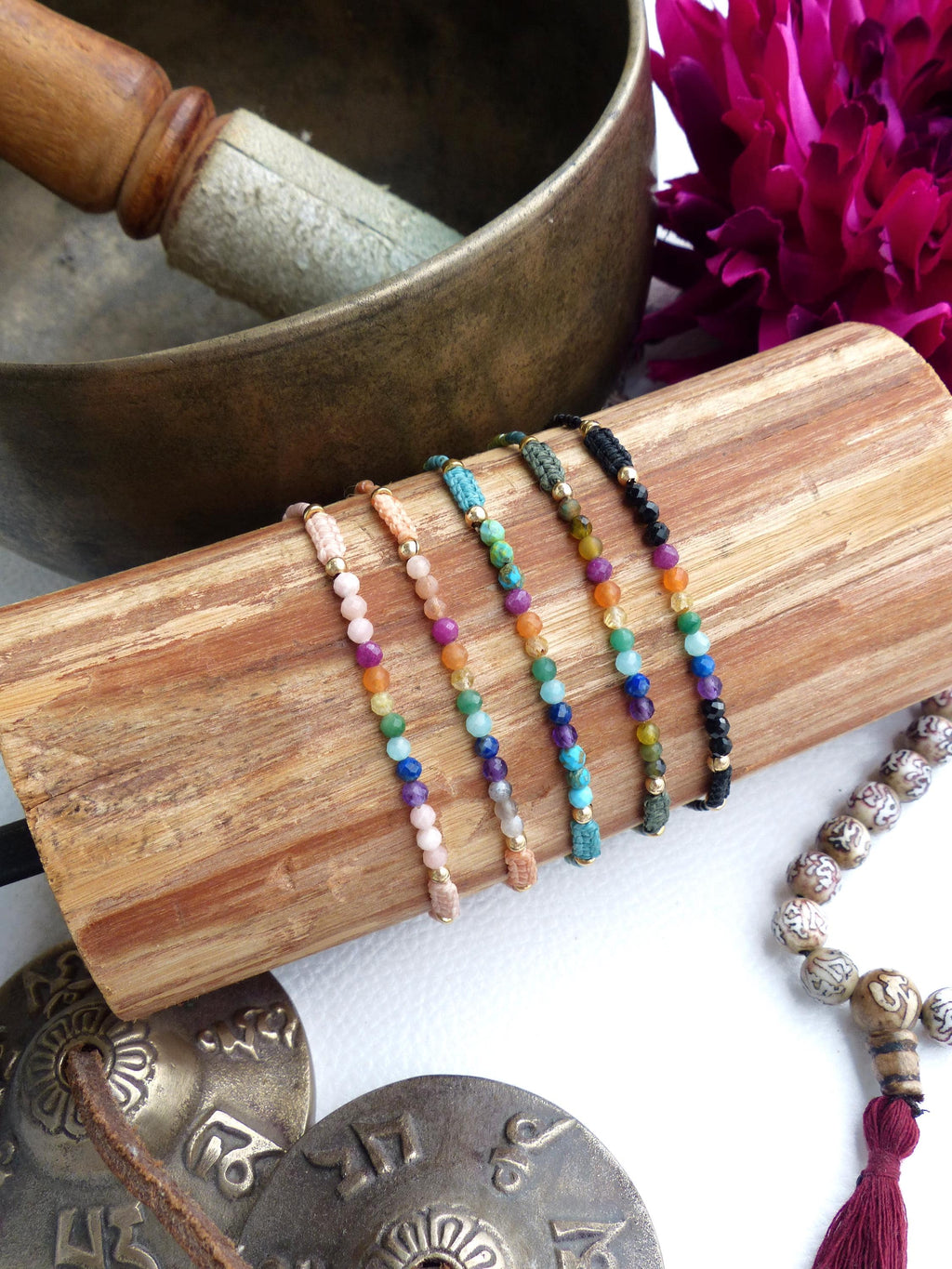 Peut inclure: Plusieurs bracelets de perles colorées, chacun avec des perles de pierres précieuses différentes, présentés sur un bloc de bois. Les bracelets comprennent des perles roses, oranges, vertes, bleues et violettes. Un bol chantant et d'autres objets spirituels sont en arrière-plan.