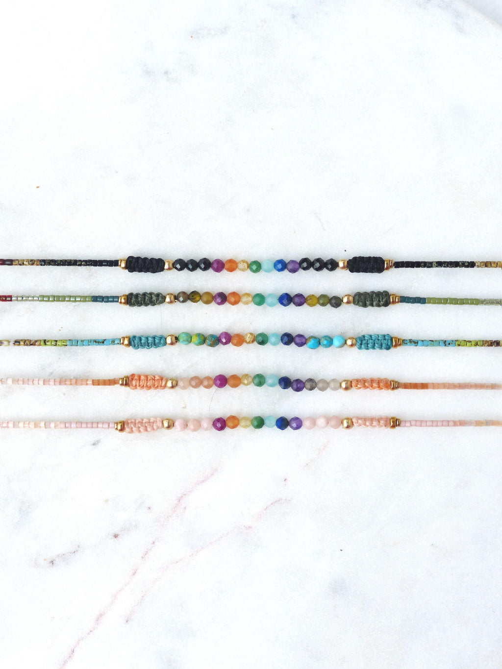 Peut inclure: Plusieurs bracelets de perles colorées. Chaque bracelet présente une rangée centrale de perles colorées, avec des détails dorés et des extrémités en cordon tressé. Les bracelets sont présentés sur une surface en marbre blanc.