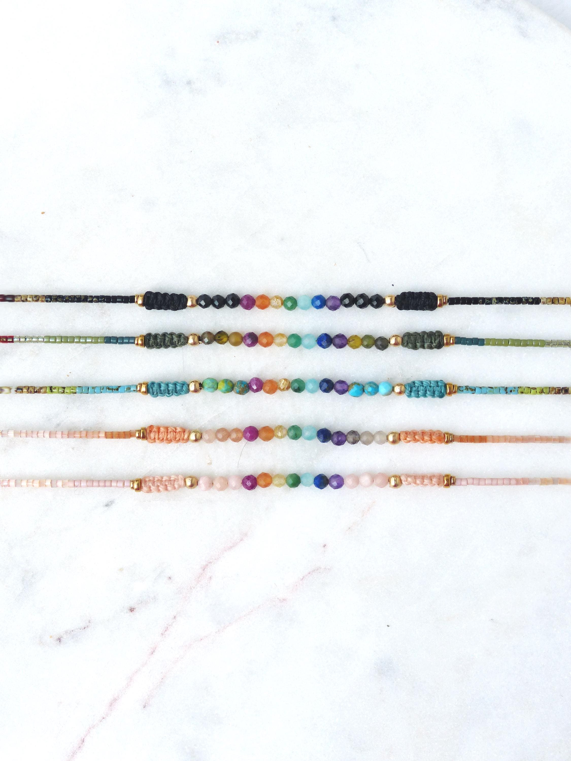 Peut inclure: Plusieurs bracelets de perles colorées. Chaque bracelet présente une rangée centrale de perles colorées, avec des détails dorés et des extrémités en cordon tressé. Les bracelets sont présentés sur une surface en marbre blanc.