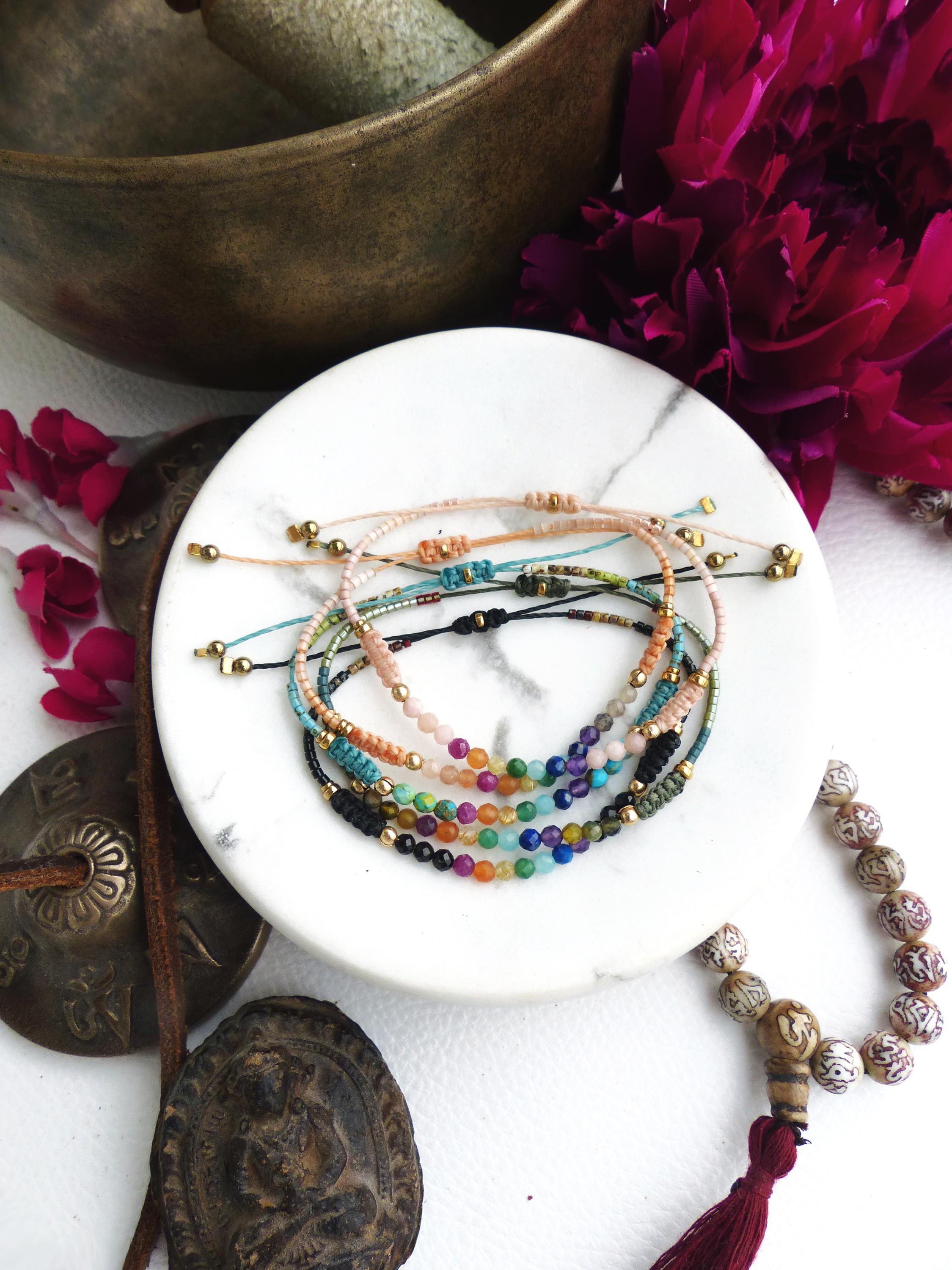 Peut inclure: Plusieurs bracelets de perles de différentes couleurs, dont rose, bleu, vert et noir, avec des détails dorés. Les bracelets sont présentés sur une surface en marbre blanc, avec un mortier en bronze et une fleur rouge foncé.